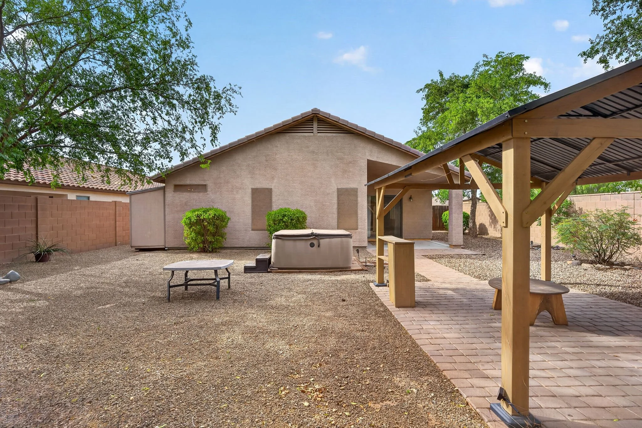 Backyard with gravel ground, a beige house with a sloped roof and closed windows, a small hot tub, a table, a shaded sitting area with a wooden picnic table, lush green trees, a brick pathway, and a brick wall. The sky is blue with a few small clouds