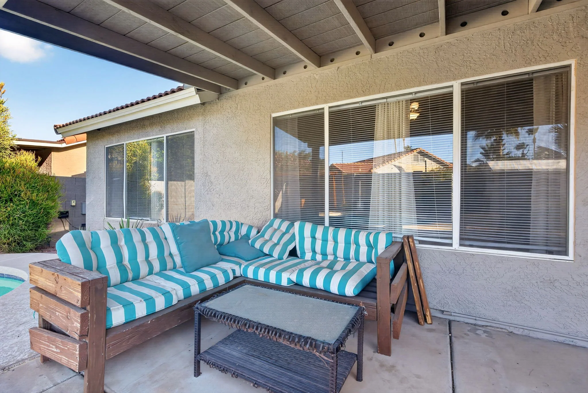 Back patio of a house showing large windows, a striped outdoor sofa, small tables, and a view of neighboring rooftops and trees.
