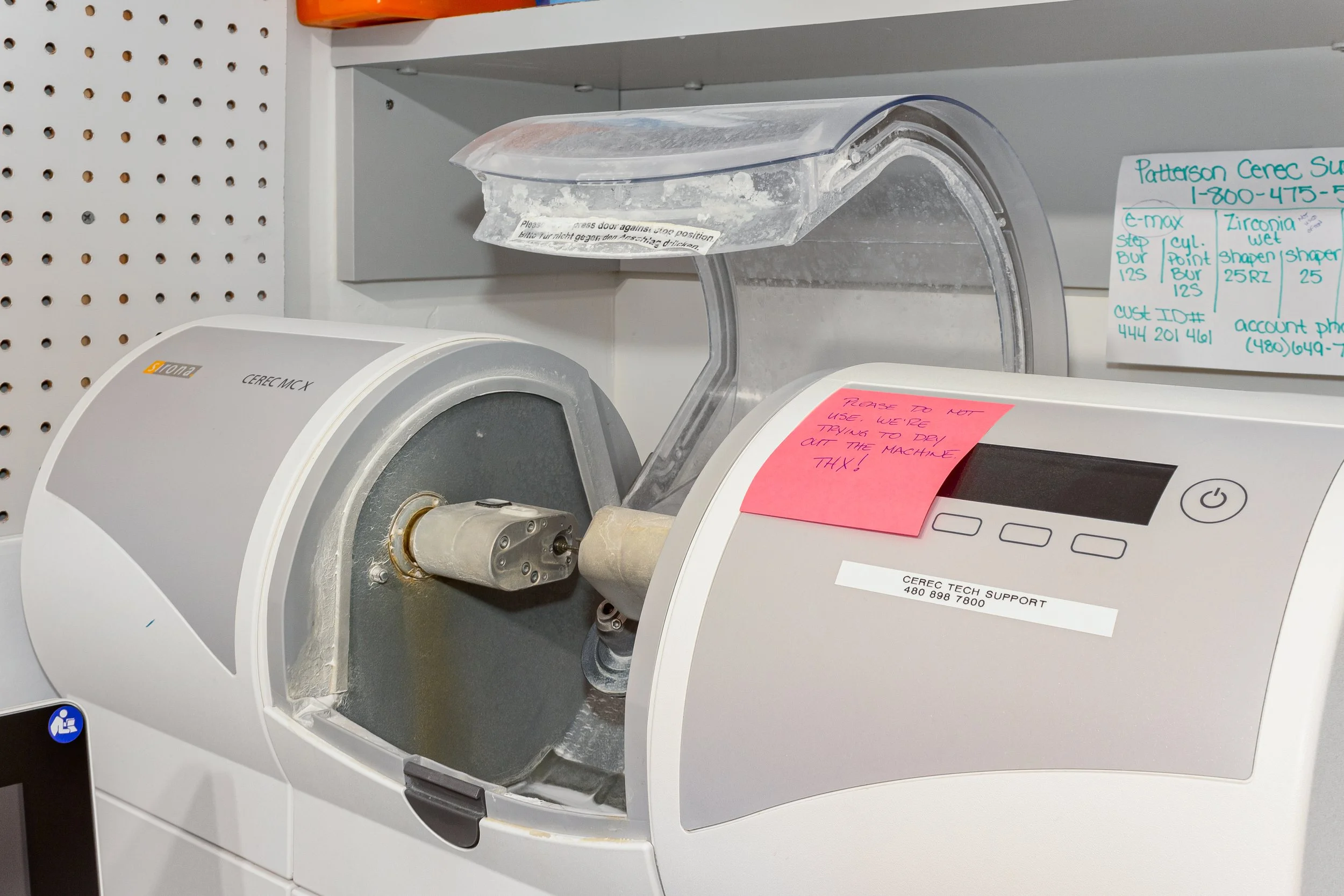 A medical centrifuge machine with a clear plastic cover, labels, and handwritten notes attached to it, placed on a shelf with a perforated white back panel.