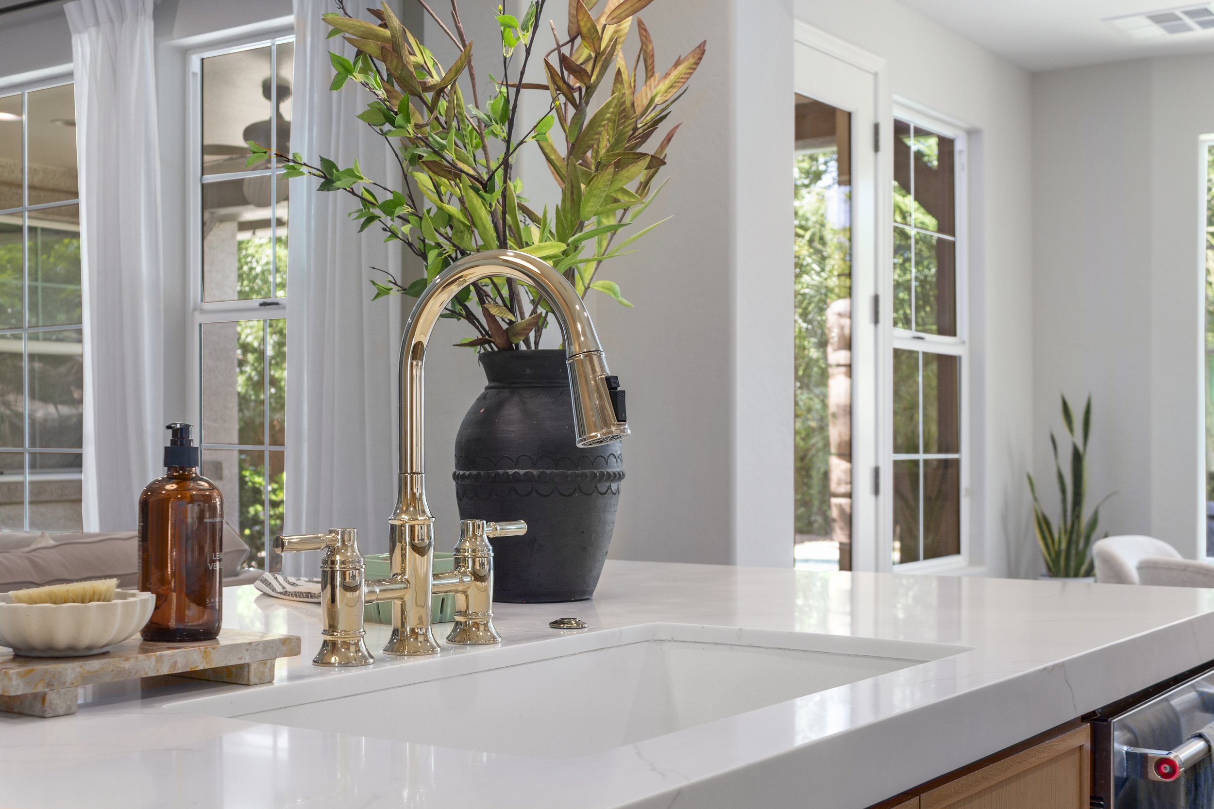A modern kitchen with a white countertop, a gold-colored faucet, a dark vase with green and brown leaves, a soap dispenser, and a small dish. Large windows with white curtains provide natural light.