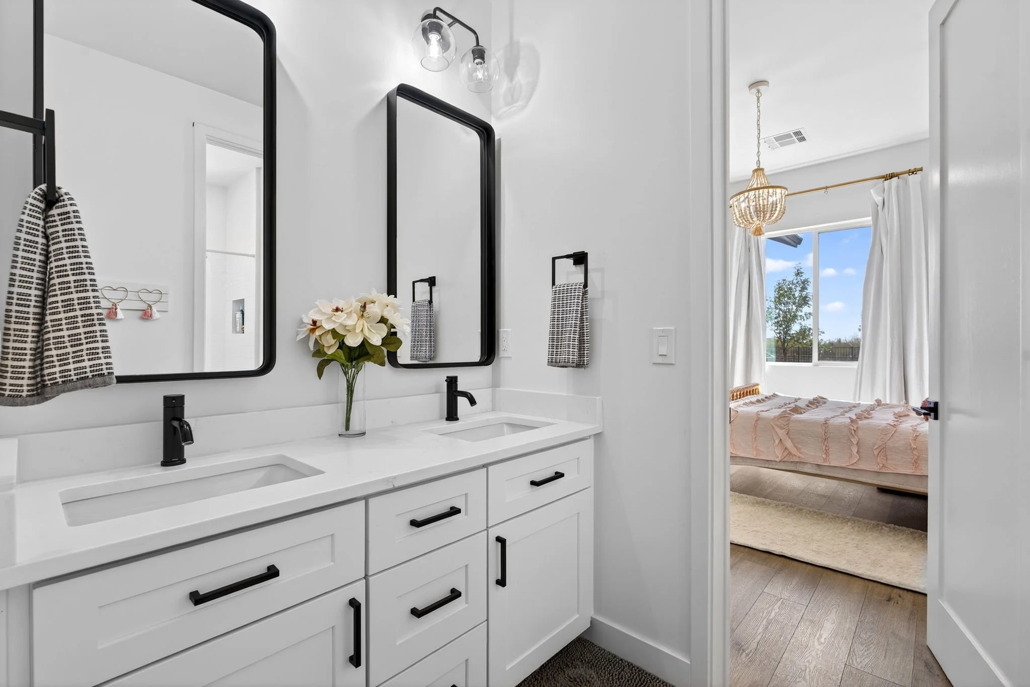 A modern bathroom with white cabinets, black handles, two black-framed mirrors, and a white countertop with two sinks. There is a vased flower on the counter and a black tap. The bathroom has a light fixture with exposed bulbs. A doorway leads to a b