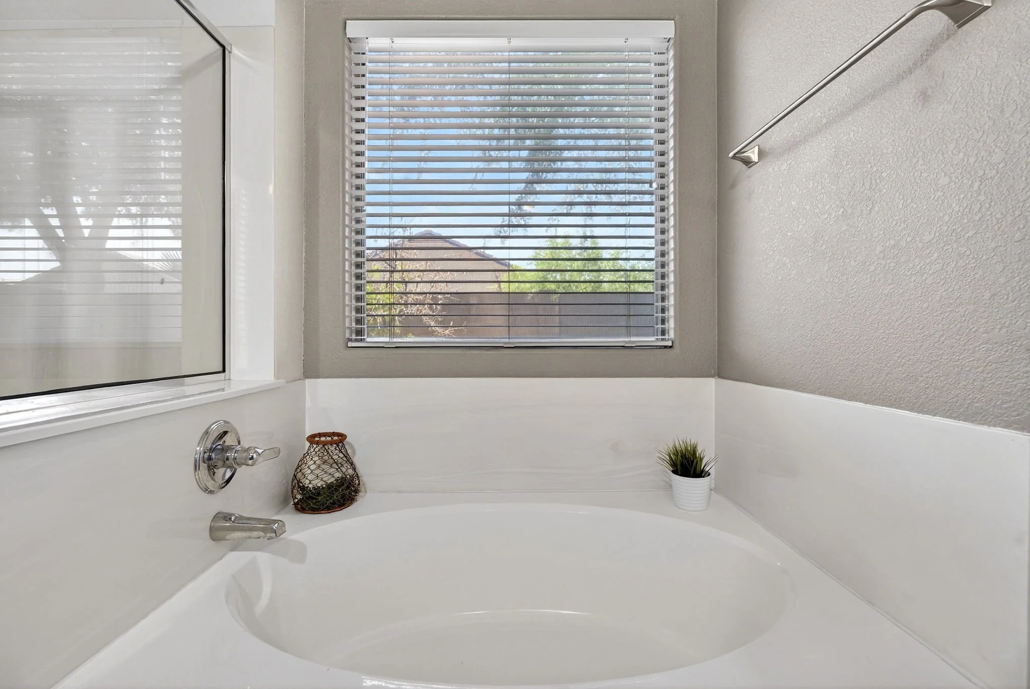 A bathroom with a white bathtub, silver faucet, and grip handle. A window with blinds shows trees outside. Decor includes a small potted green plant and a decorative wire basket with plants inside.