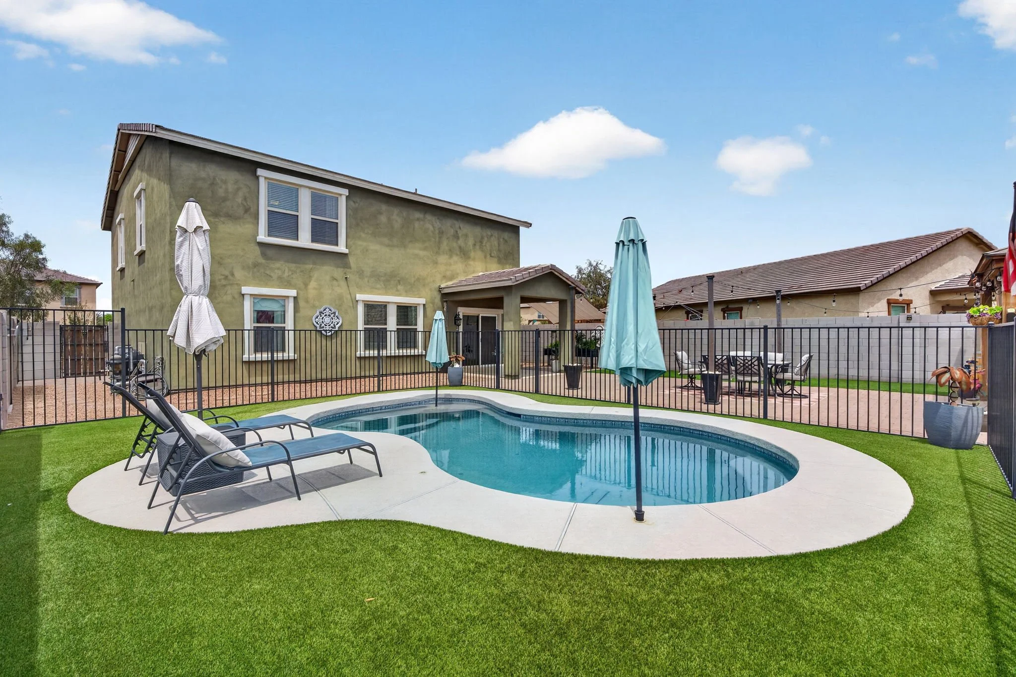 Backyard with a swimming pool, lounge chairs, umbrellas, patio furniture, a green two-story house, and a blue sky with clouds.