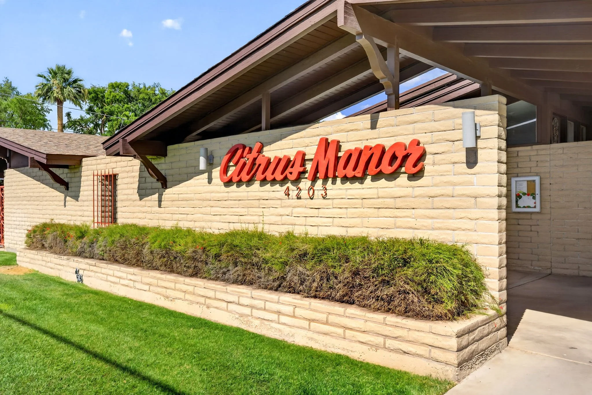 The image shows the exterior of a building with a brick wall and a sign reading 'Tulsa Manor' in red cursive letters, with the numbers 4203 underneath, surrounded by greenery including a small shrub and a tall palm tree in the background.