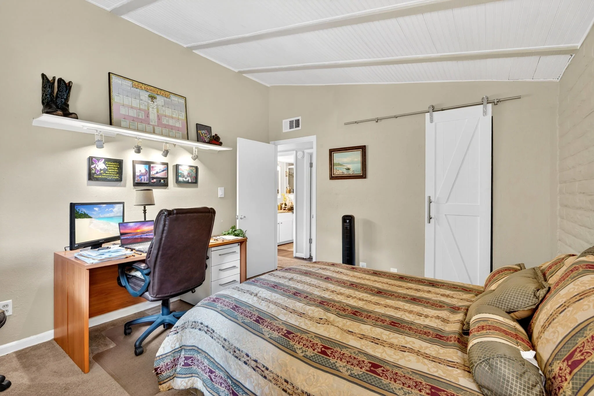 A bedroom with a bed, computer desk, office chair, framed artwork, and decorative items on shelves, with a door leading to another room.