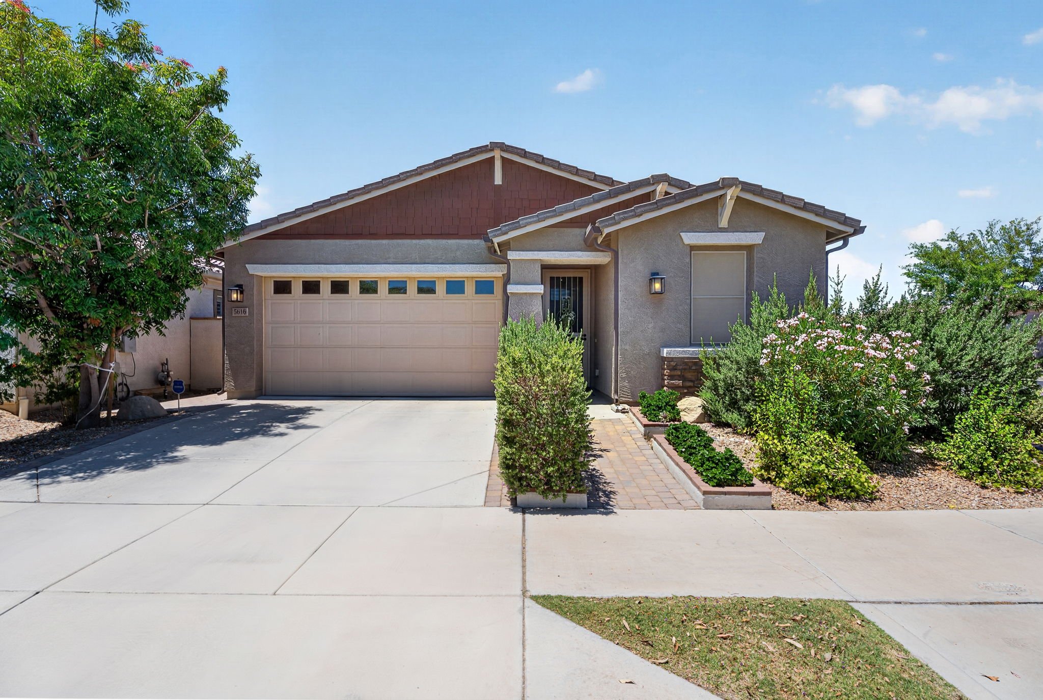 Front view of a house with a two-car garage, driveway, and landscaped front yard with bushes and flowering plants, under a blue sky with a few clouds.