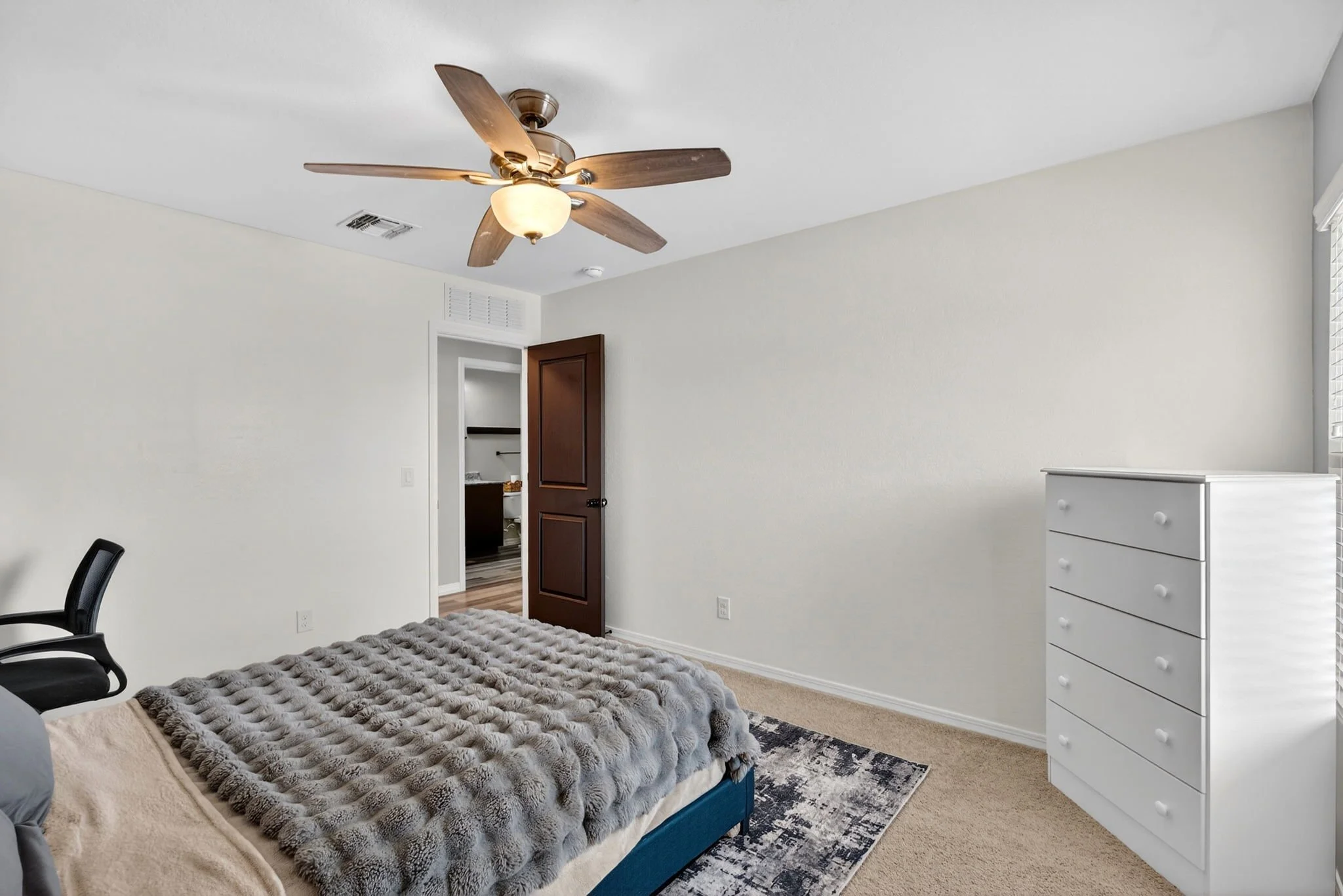 A bedroom with a bed with a textured gray blanket, black chairs, a white dresser, and a ceiling fan with wooden blades and a light. The room has cream walls, a window with blinds, and an open door leading to a bathroom.