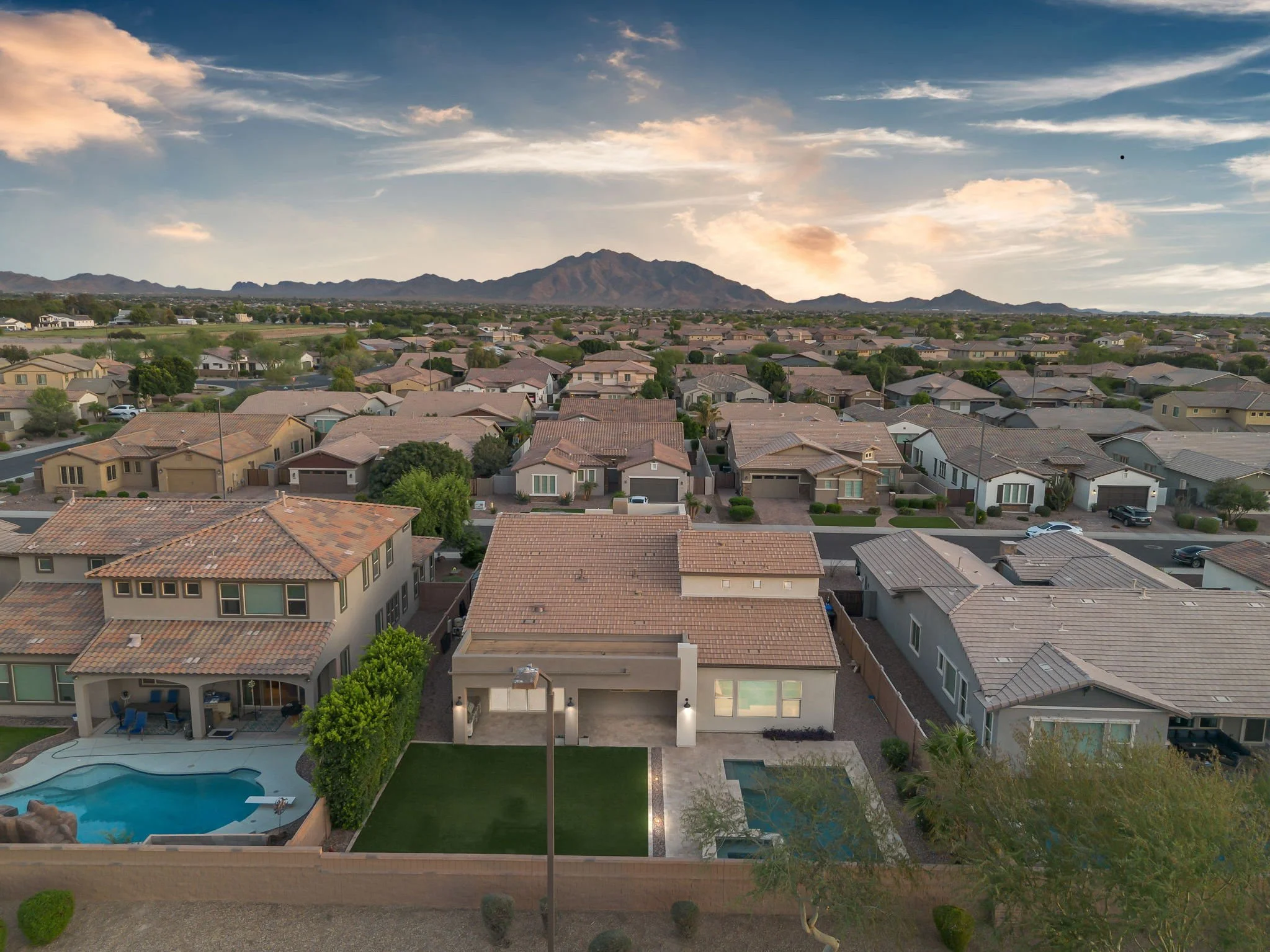 Aerial view of a suburban neighborhood with single-family homes, a swimming pool, and a backyard with a patio and grass, set against mountains in the distance under a partly cloudy sky.