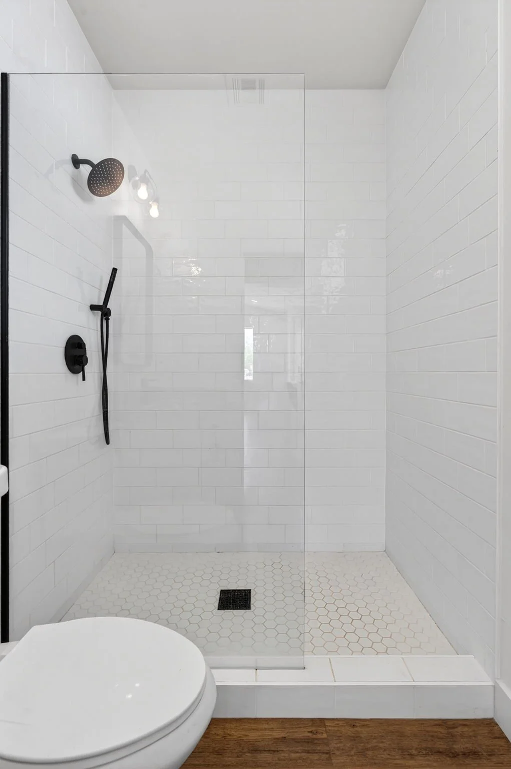 Modern bathroom shower with black fixtures, glass door, white subway tiles, and a white toilet in the foreground, with wooden flooring outside the shower.