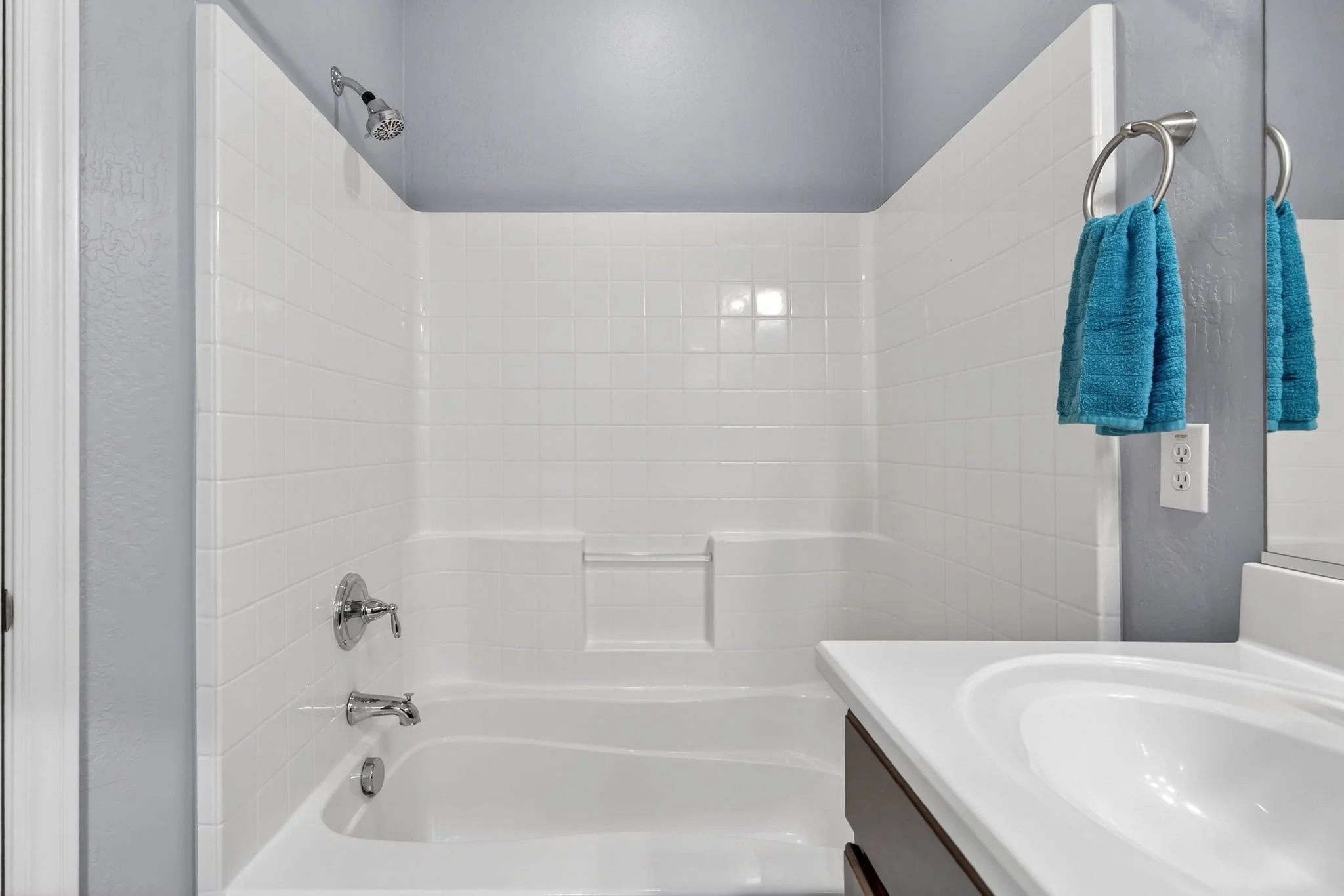 A white bathtub with chrome fixtures, two blue towels hanging on hooks, and white tiled walls in a bathroom.