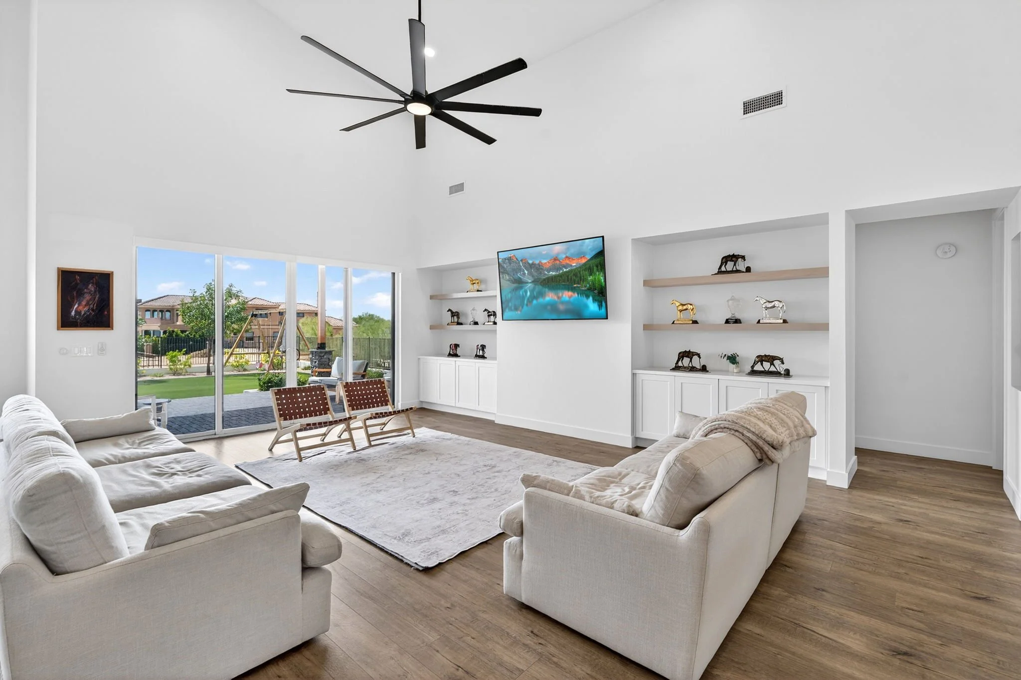 Bright living room with white sofas, a large rug, wooden floor, and a wall-mounted TV showing a mountain landscape. Windows show a backyard with trees and a patio.