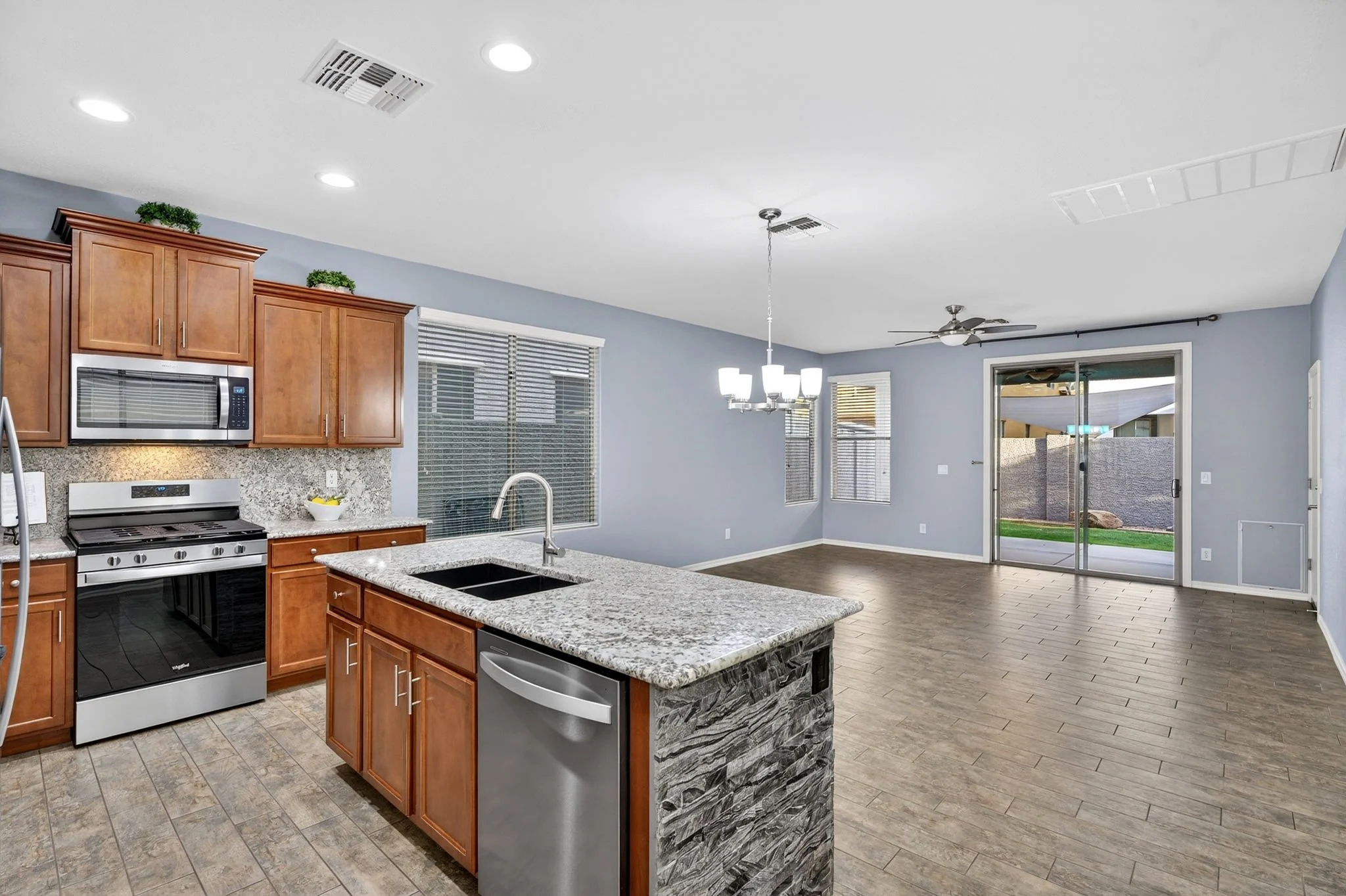 A modern open-concept kitchen and living area with wooden cabinets, a granite island, stainless steel appliances, large windows, a ceiling fan, and a sliding glass door leading to an outdoor space.