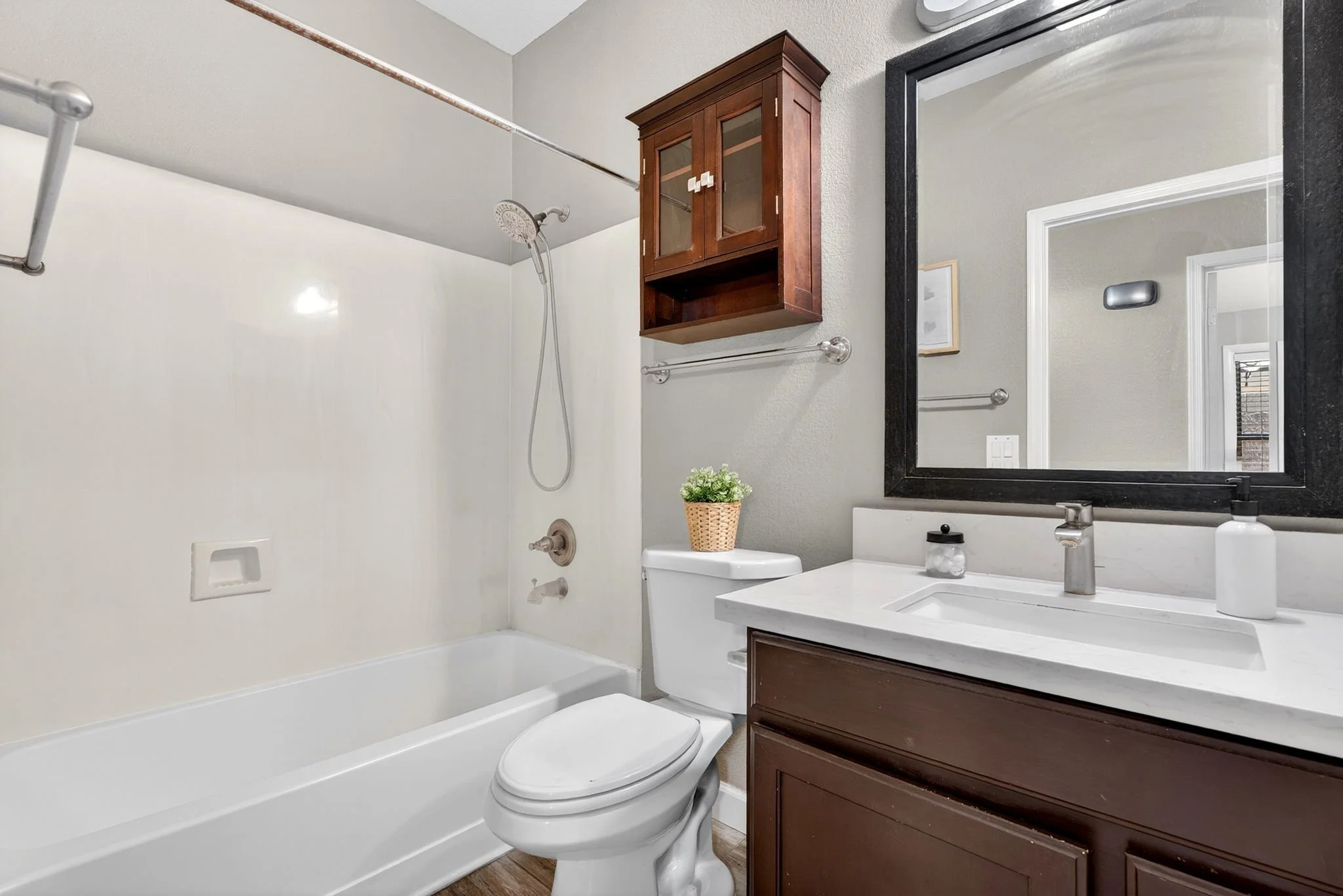 Bathroom with bathtub, showerhead, toilet, wooden cabinet above the toilet, large mirror, sink with soap dispenser, and small plant on the toilet tank.