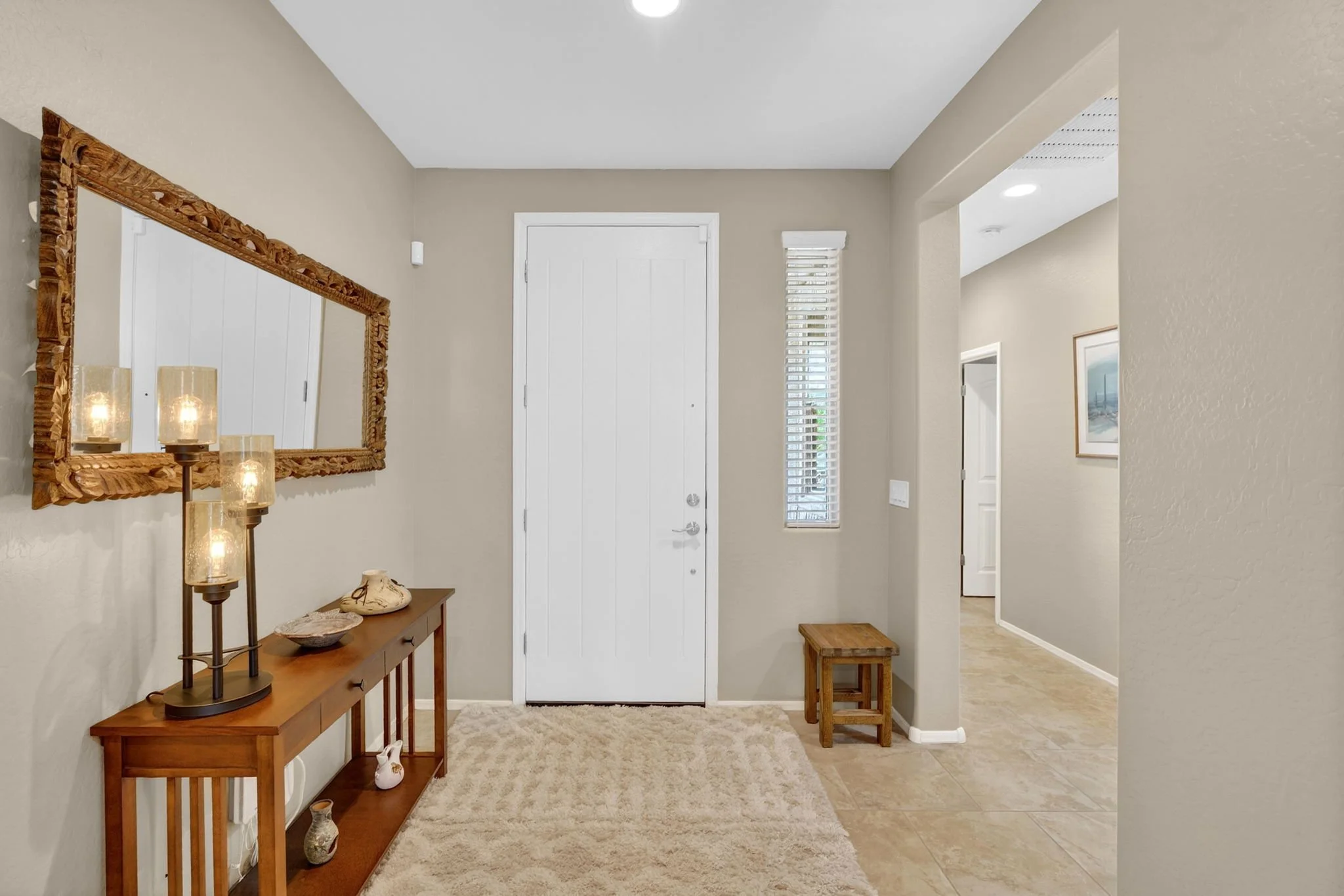 Entranceway with a white front door, a wooden console table with decorative items, wall-mounted lighting, a large wall mirror, and a small wooden stool, with beige walls and tile flooring.