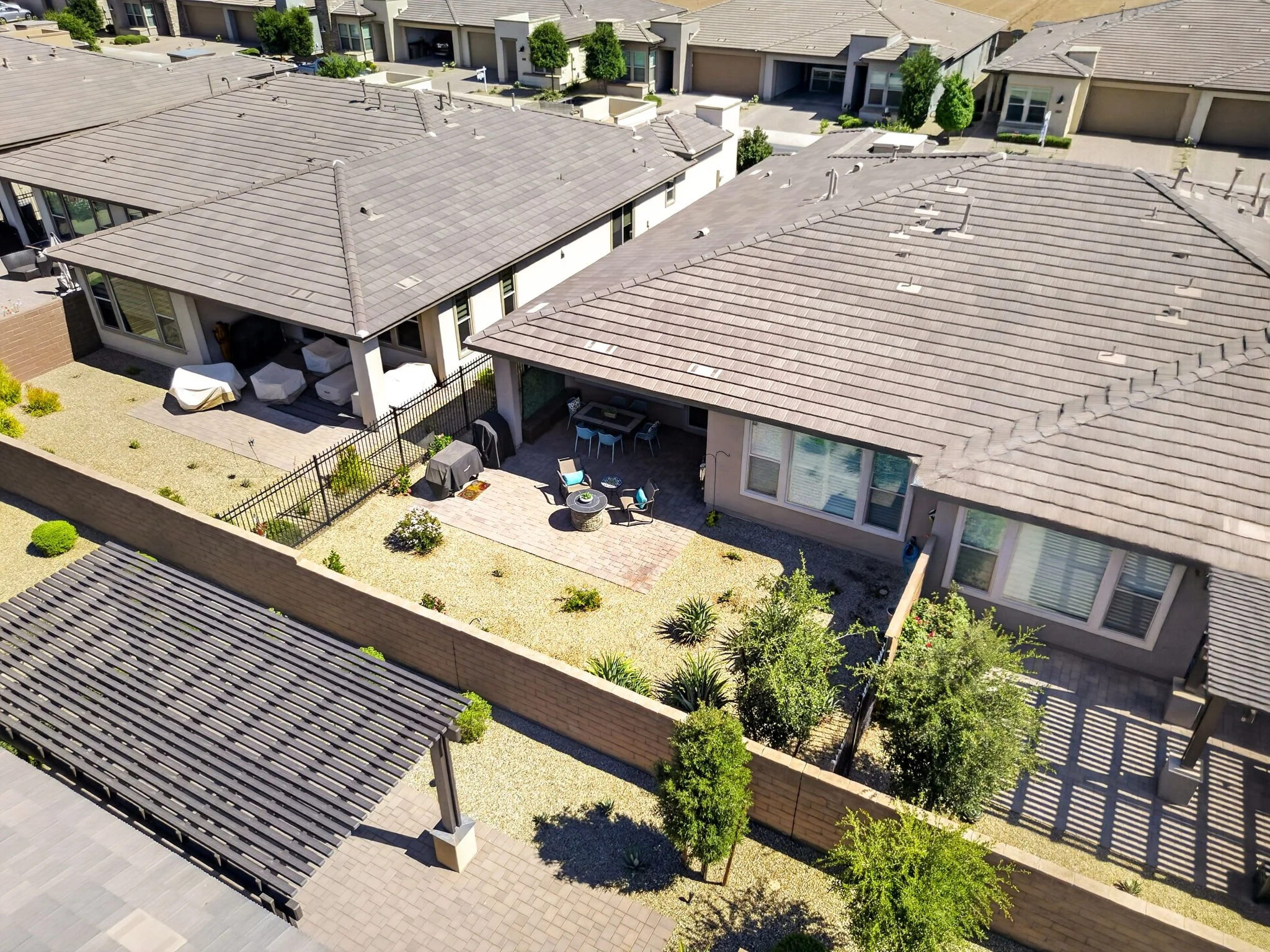 Aerial view of a residential neighborhood with multiple houses, gray tiled roofs, backyard patios with outdoor furniture, and landscaped gardens.