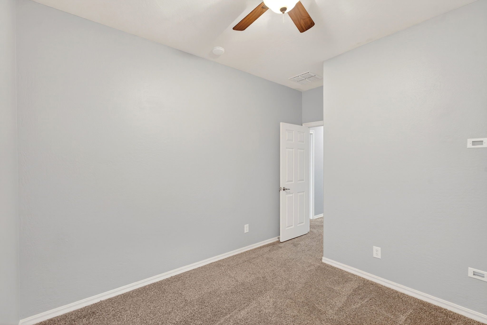 An empty room with light gray walls, a ceiling fan with wooden blades, a smoke detector, a white door partially open, a thermostat on the wall, and beige carpet flooring.