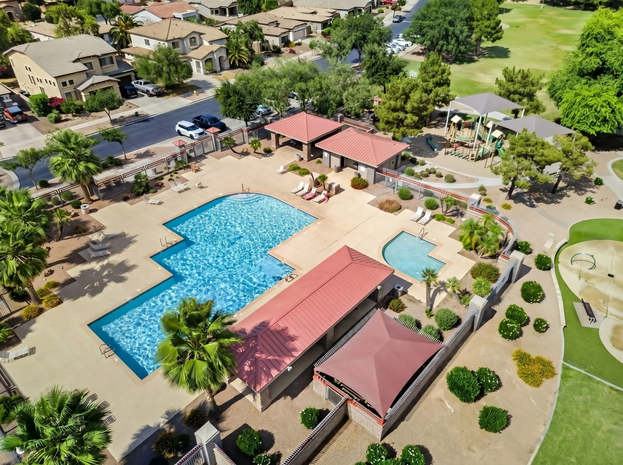 An aerial view of a community pool area surrounded by a fence, with lounge chairs, palm trees, and shaded structures. There are nearby residential houses and a green park area with trees and a playground.
