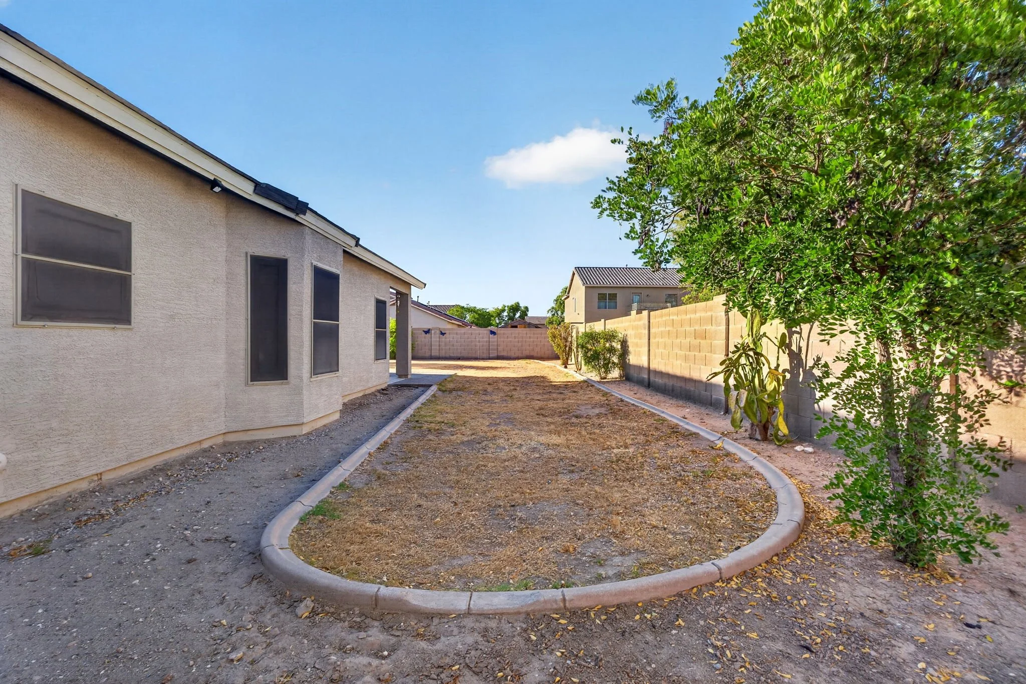 Backyard with a single-story house, a tree on the right, a brick wall fence, and a patch of dirt and gravel ground.