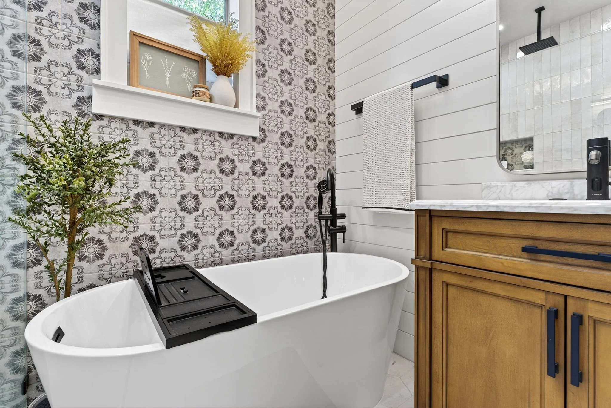 Modern bathroom with a white bathtub, black faucet, white shiplap walls, patterned tile, wooden cabinet, mirror, potted plant, window with floral art, shelves with decorative items, and a towel rack.