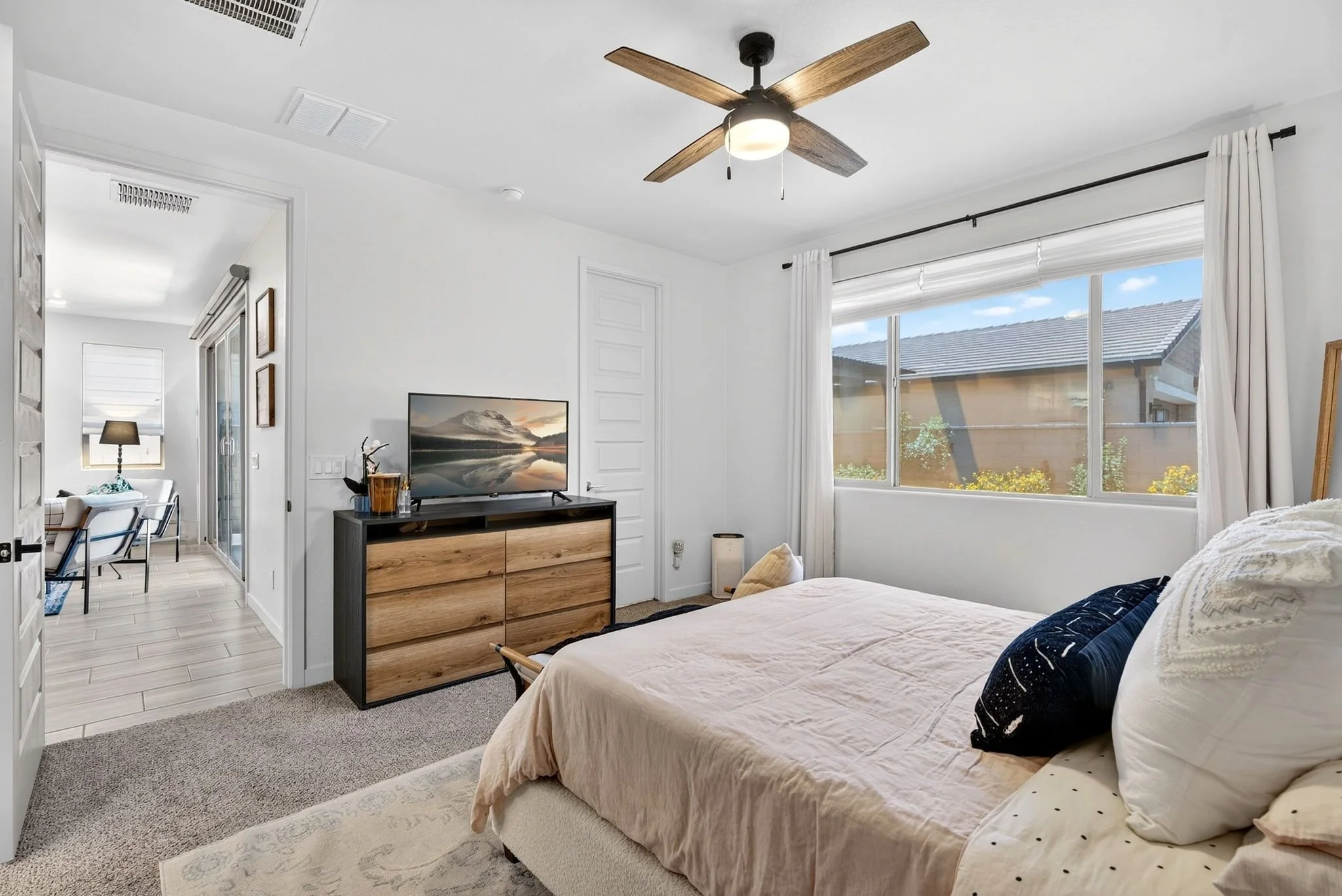 A bedroom with a large window, beige bed with pillows, a black and wood dresser with a TV, and a ceiling fan with wooden blades.