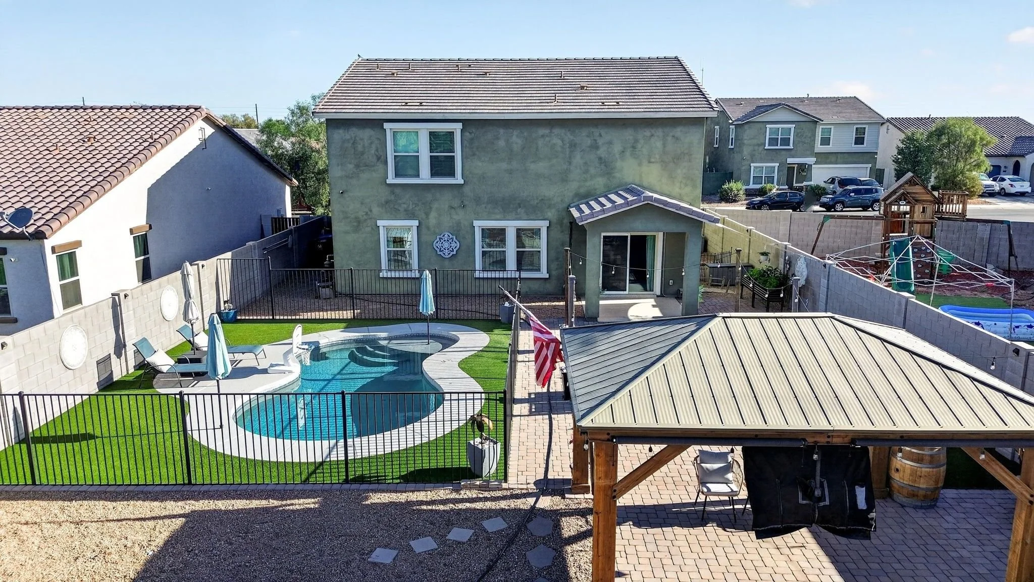 A backyard with a swimming pool, poolside chairs, and umbrellas, enclosed by a black metal fence, with a house painted in green and neighboring houses in the background.