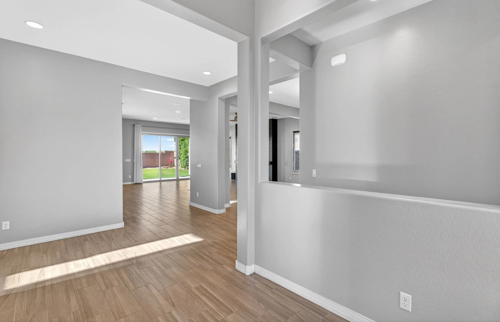 An interior view of a modern, minimalistic living space with white walls, hardwood flooring, and large sliding glass doors leading to a backyard with greenery and a patio.