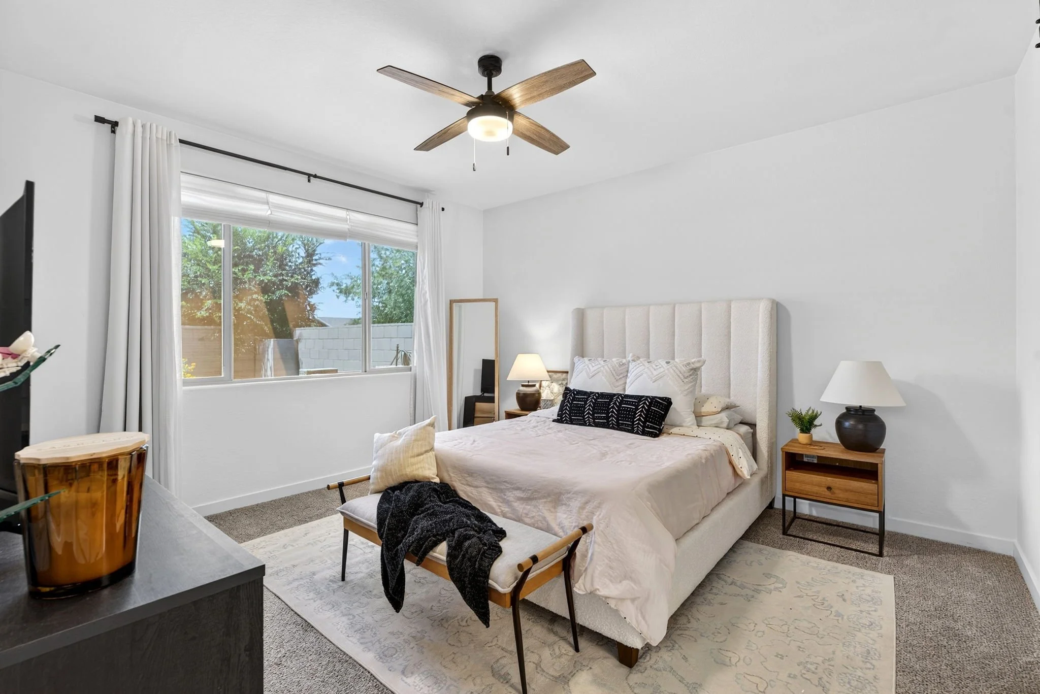 A cozy bedroom with a large window, white walls, and a beige upholstered bed with multiple pillows. There are two nightstands with lamps, a full-length mirror, and a ceiling fan with wooden blades.
