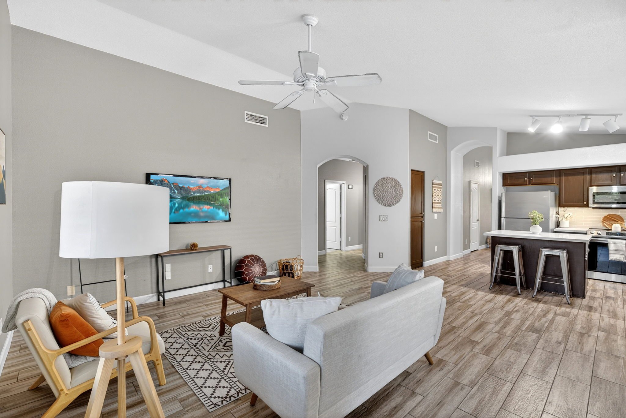 Open-concept living room and kitchen with gray walls, wooden floors, modern furniture, a hanging ceiling fan, a mounted TV, and kitchen cabinets.