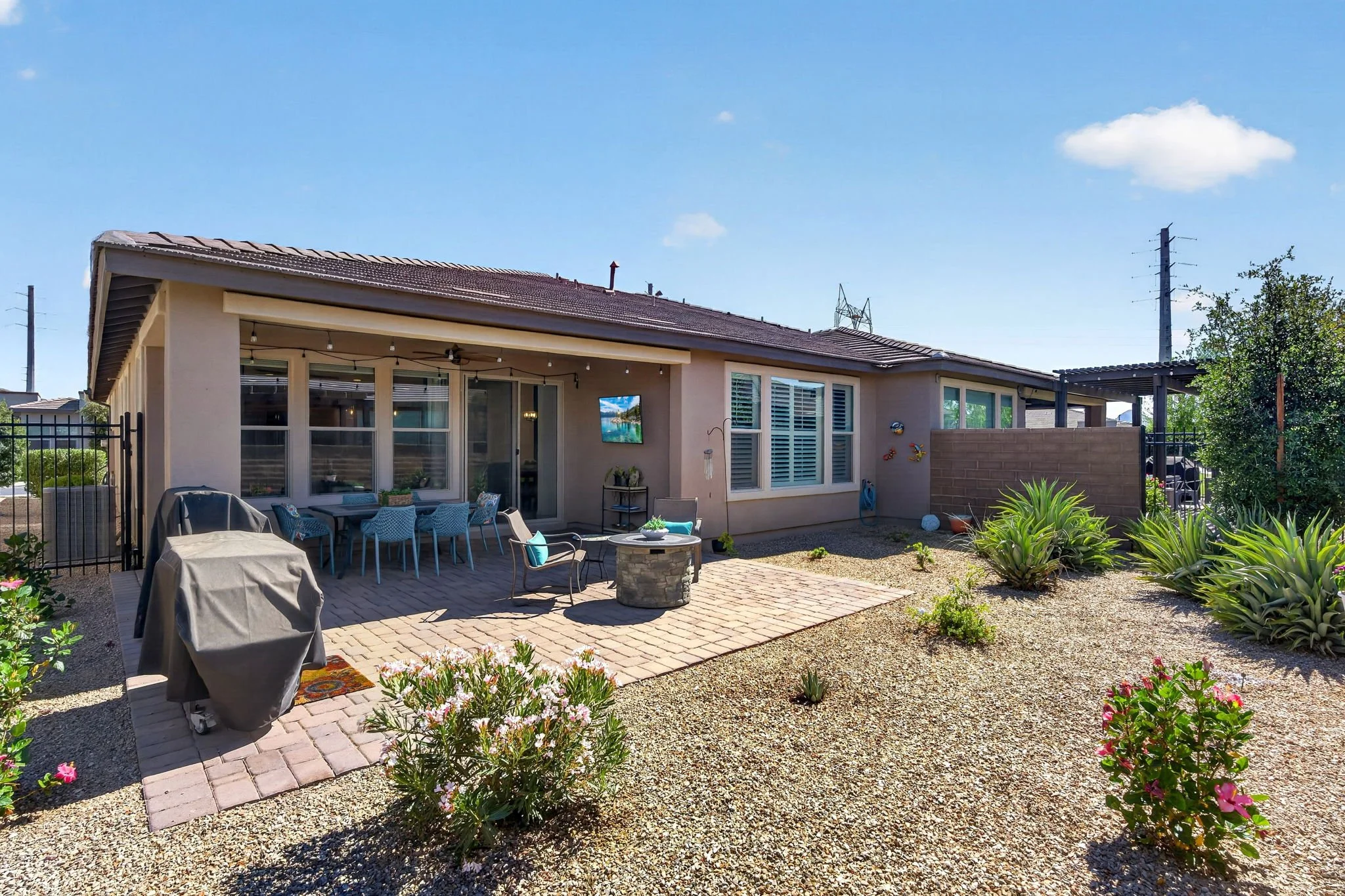 Backyard patio with outdoor dining area, a grill covered with a cloth, and a flower bed with blooming plants, adjacent to a house with large windows and a backyard garden.
