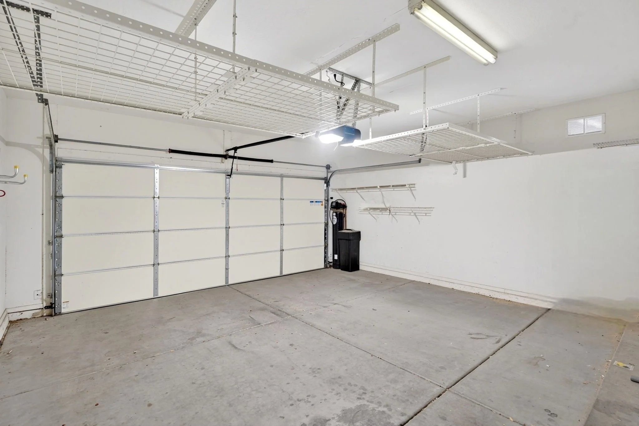 Empty garage with concrete floor, white walls, and a closed white garage door. Overhead lighting and wall-mounted racks are visible.