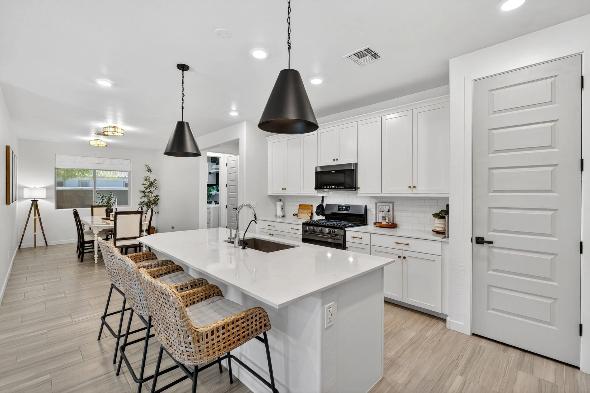 A modern kitchen and dining area with white cabinets, black pendant lights, and a white kitchen island with wicker chairs. The dining area has a table with black chairs, a floor lamp, and a window with a view outside.