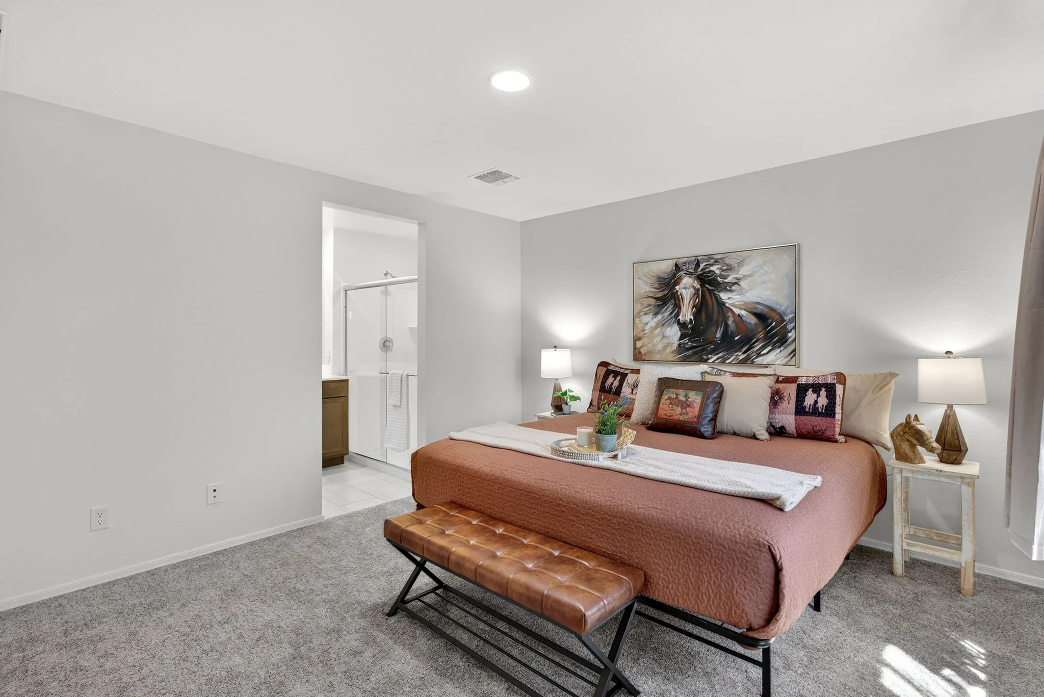 A bedroom with a bed covered in a pinkish bedspread, decorated with pillows, a plant, and a tray with candles on a bench at the foot of the bed. There are two nightstands with lamps and artwork of a horse above the bed. The room has gray carpet and w