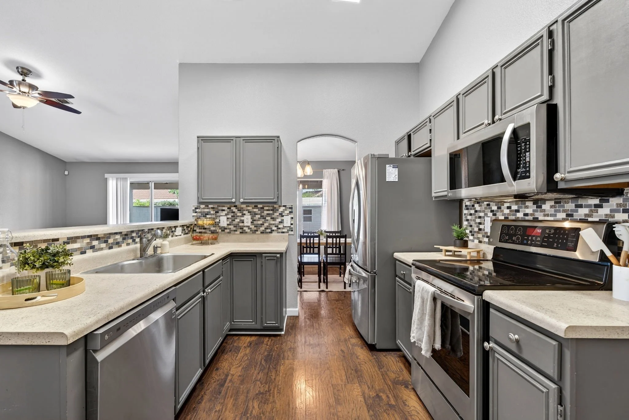 A kitchen with gray cabinets, a white countertop, and a mosaic tile backsplash. There is a stainless steel microwave above a stove, a refrigerator, and a small dining area with a table and four chairs in the background. The kitchen has wood flooring,