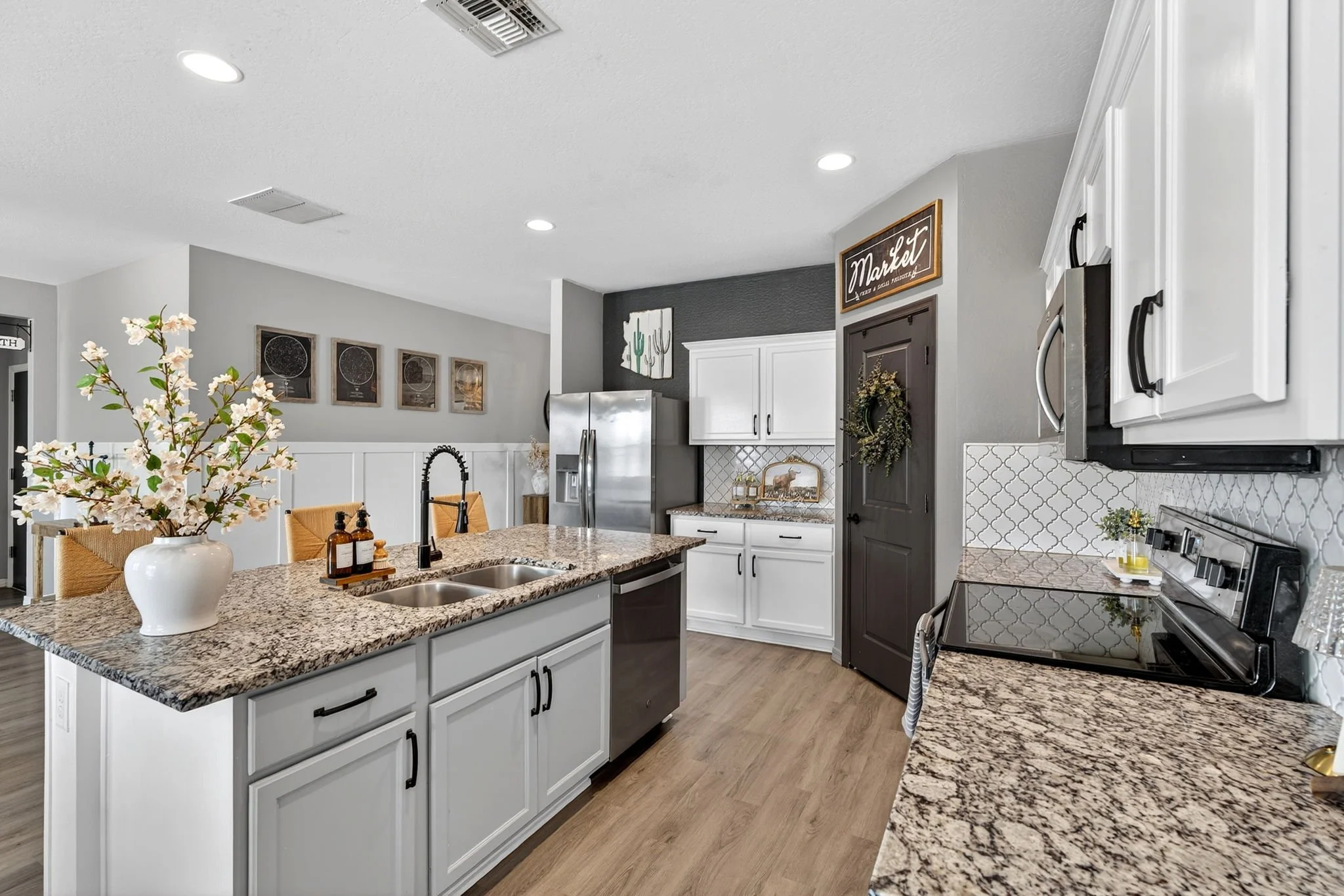 Modern kitchen with white cabinets, granite countertops, a kitchen island, stainless steel refrigerator, black stove, white shiplap wainscoting, decorative wall art, and a vase with flowers on the island.