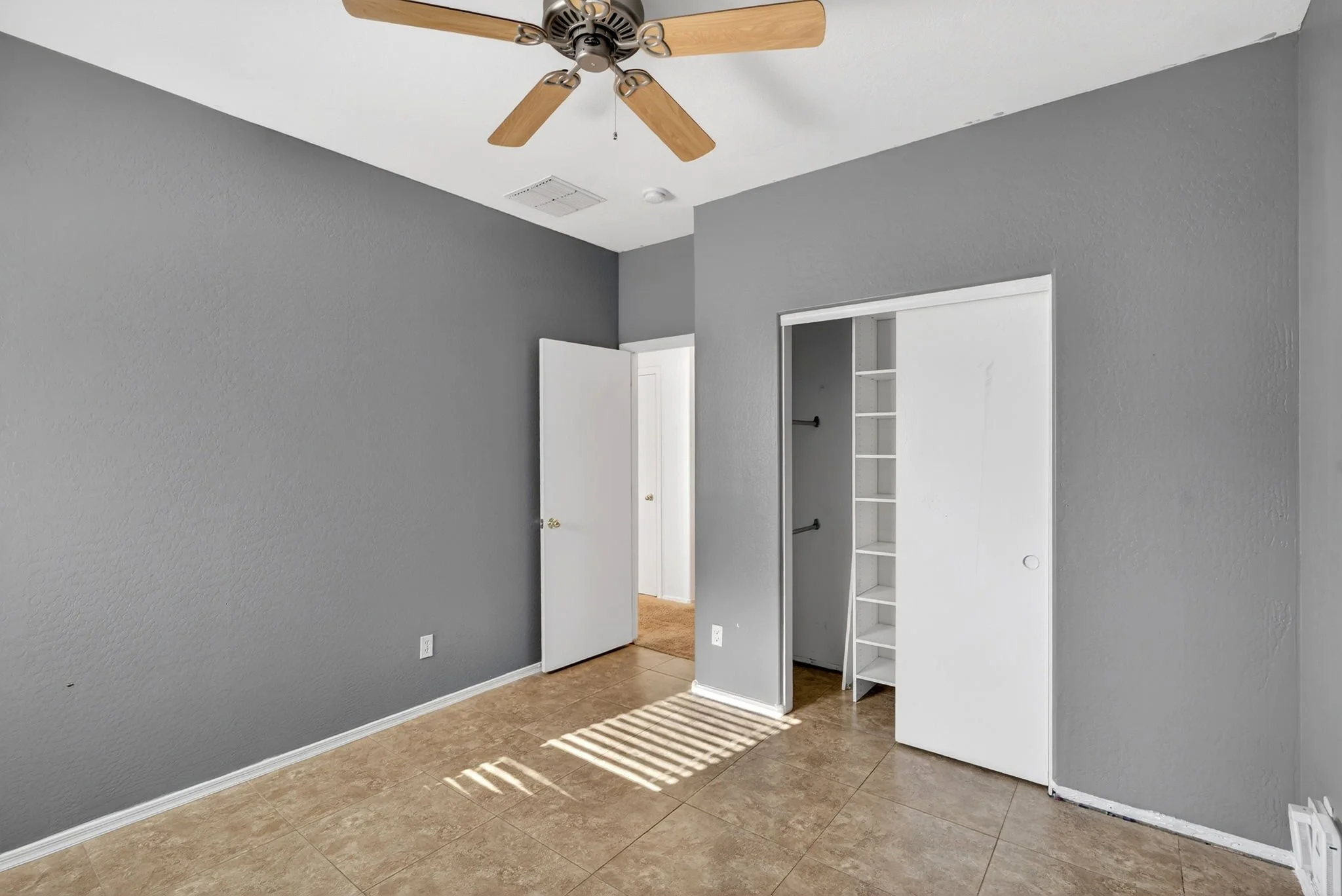 Empty room with gray walls, a closet with open doors, a ceiling fan, and sunlight casting shadows on the tiled floor.