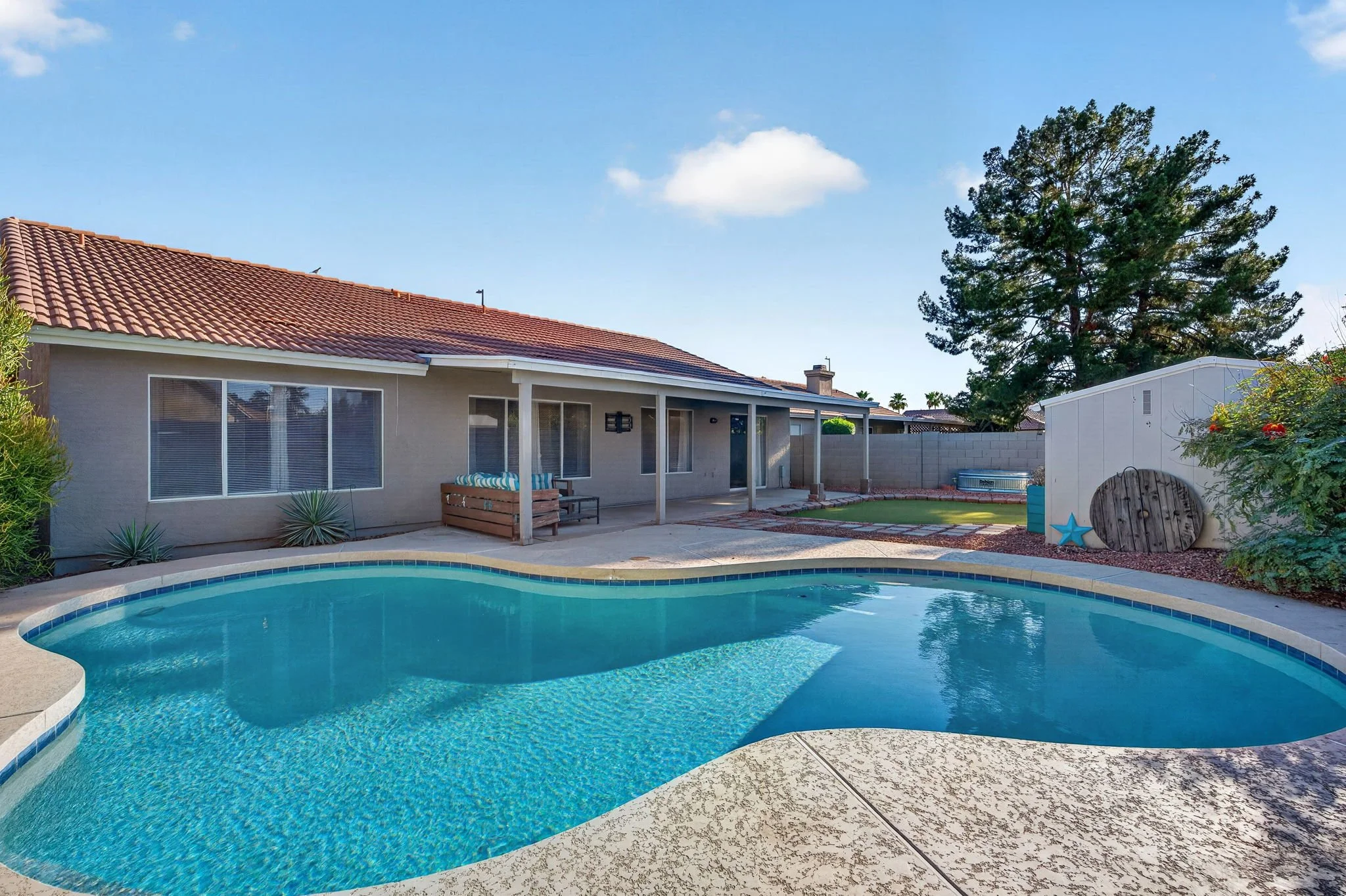 Backyard with a swimming pool, patio area, and house with a red-tiled roof, large windows, and a tree in the background.