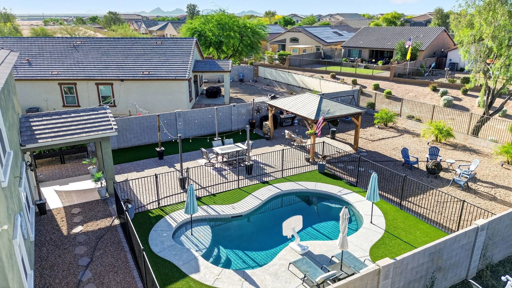 Backyard with swimming pool, patio furniture, gazebo, and fenced yard in suburban neighborhood with houses and mountains in the background.