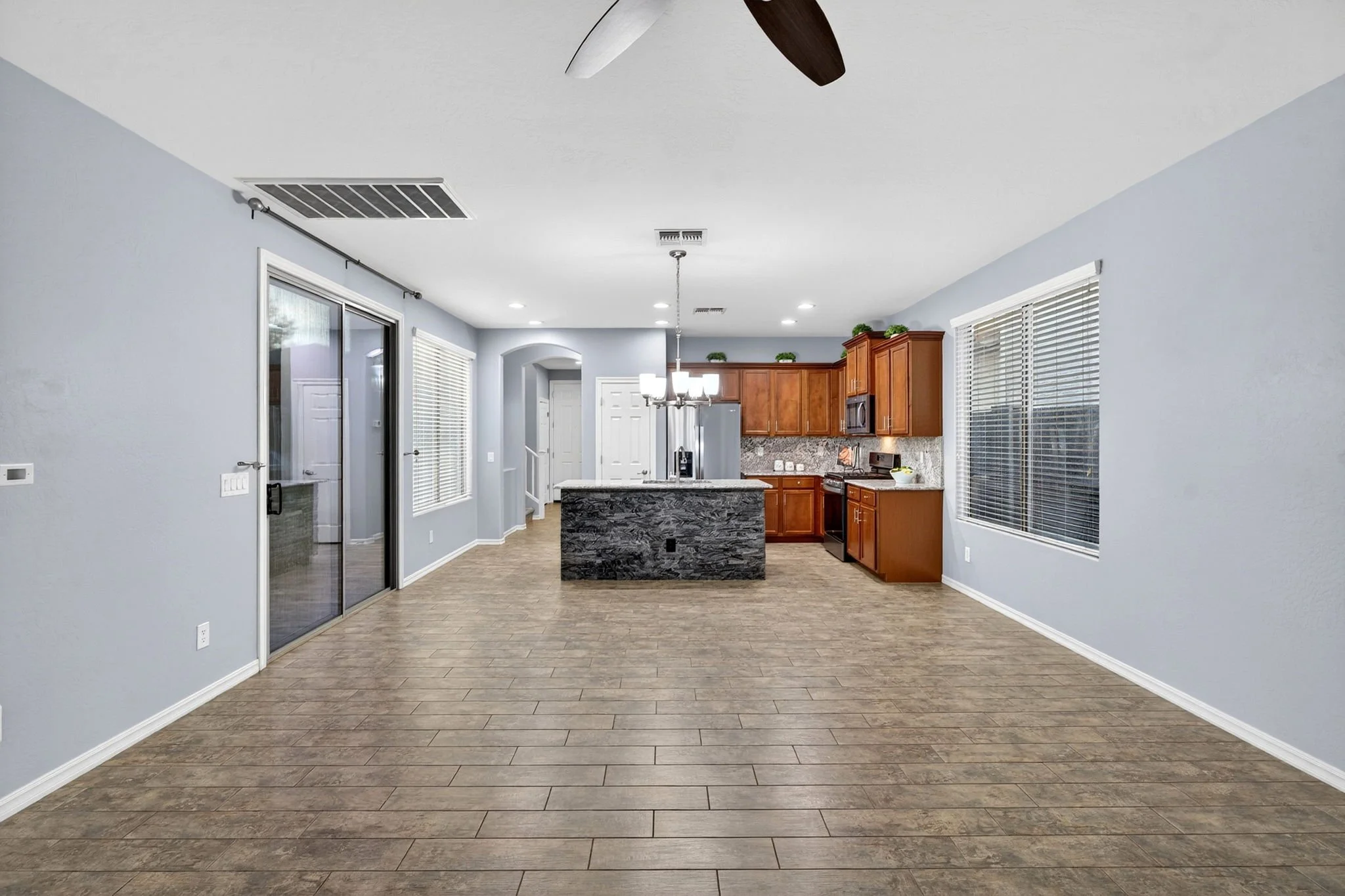 Empty living room with light blue walls, hardwood floors, large windows with blinds, a kitchen with wooden cabinets, a granite island, and modern appliances, under a ceiling fan and recessed lighting.