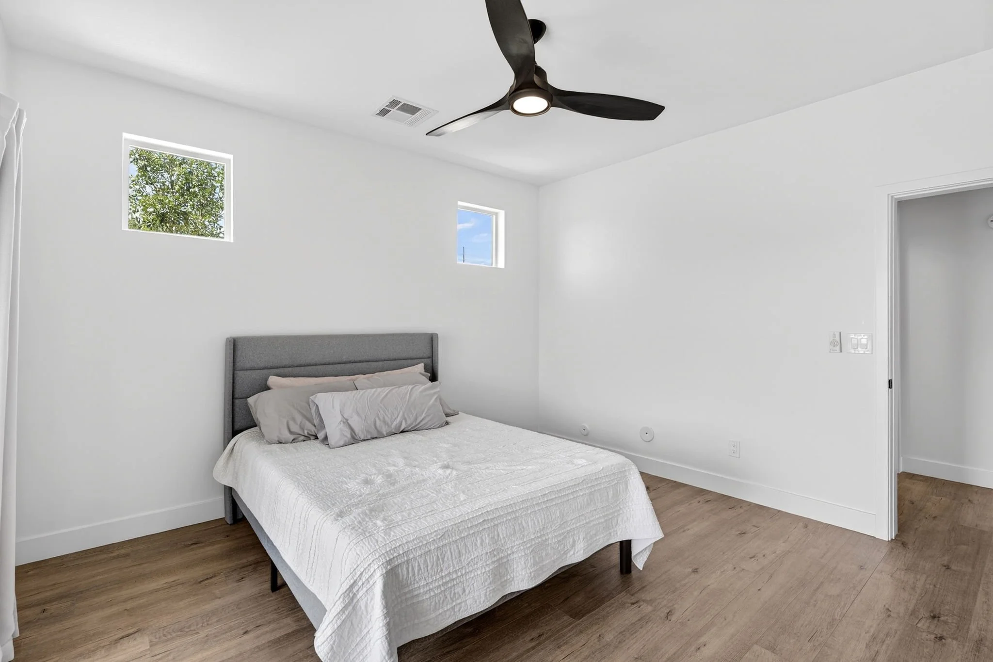 A bedroom with white walls, a bed with gray headboard and white bedding, two small rectangular windows, a ceiling fan with black blades and light, and wood flooring.