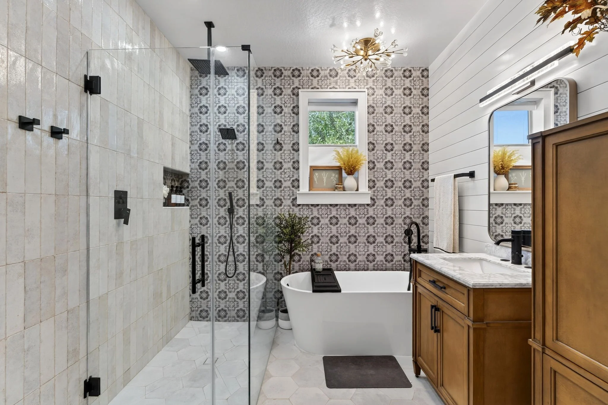 Bathroom with a walk-in shower on the left, a bathtub in the center, and a wooden vanity with a marble countertop on the right. The wall behind the bathtub has floral patterned tiles, and there is a small window with a vase of flowers on the windowsi