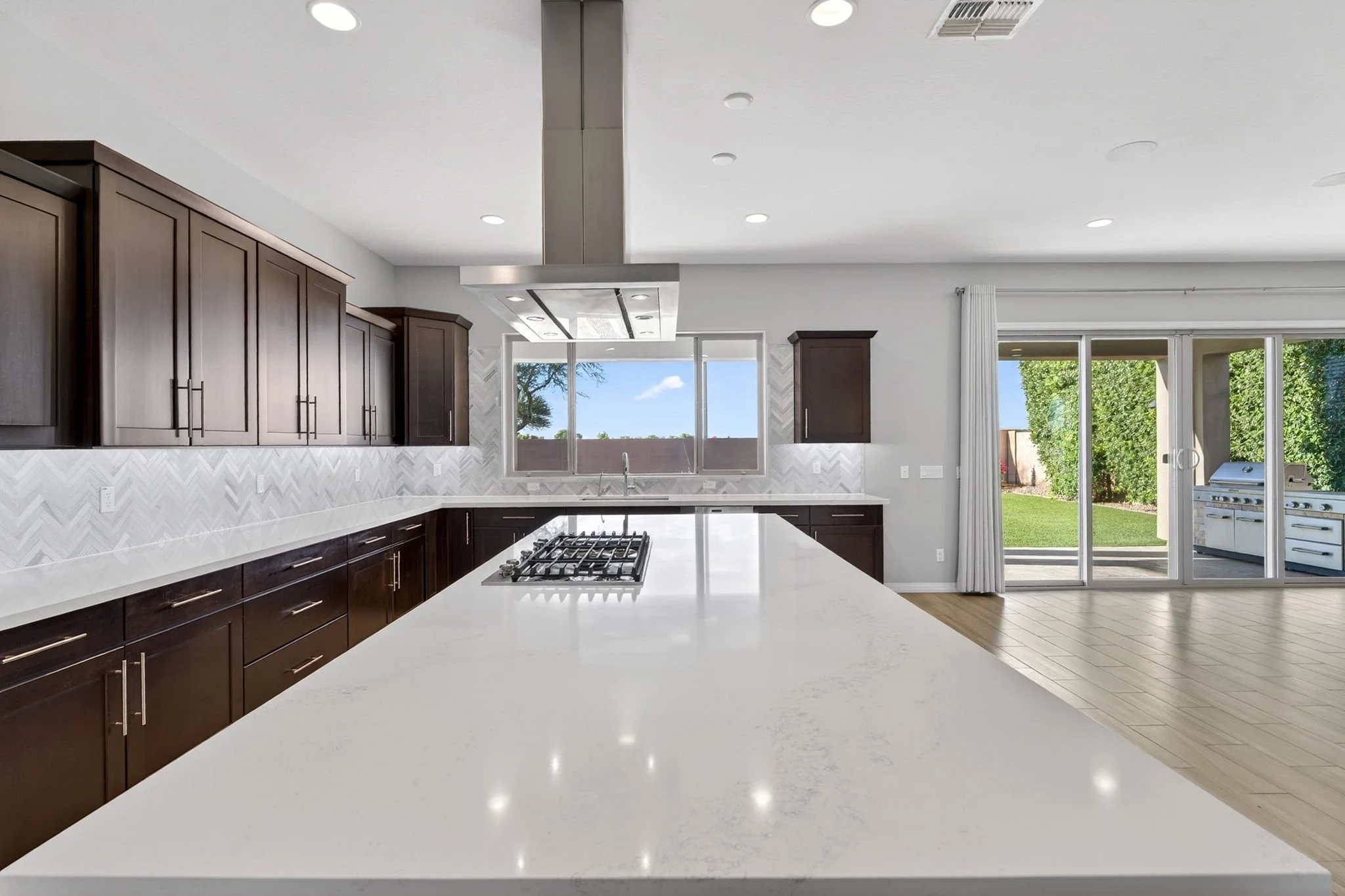A modern kitchen with dark wooden cabinets, white countertops, a central island, and large windows that overlook a backyard with a grill and trees.