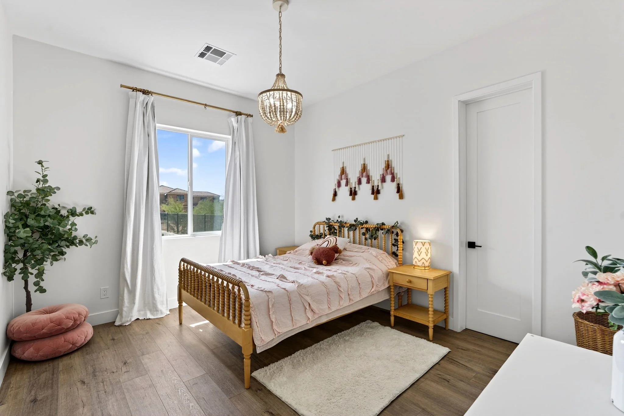 A bedroom with a wooden bed, pink striped bedding, and a decorative pillow. A nightstand with a lamp and a plant is beside the bed. There are white curtains over a window, a wall hanging with tassels, a potted plant, and a pink quilted floor cushion.