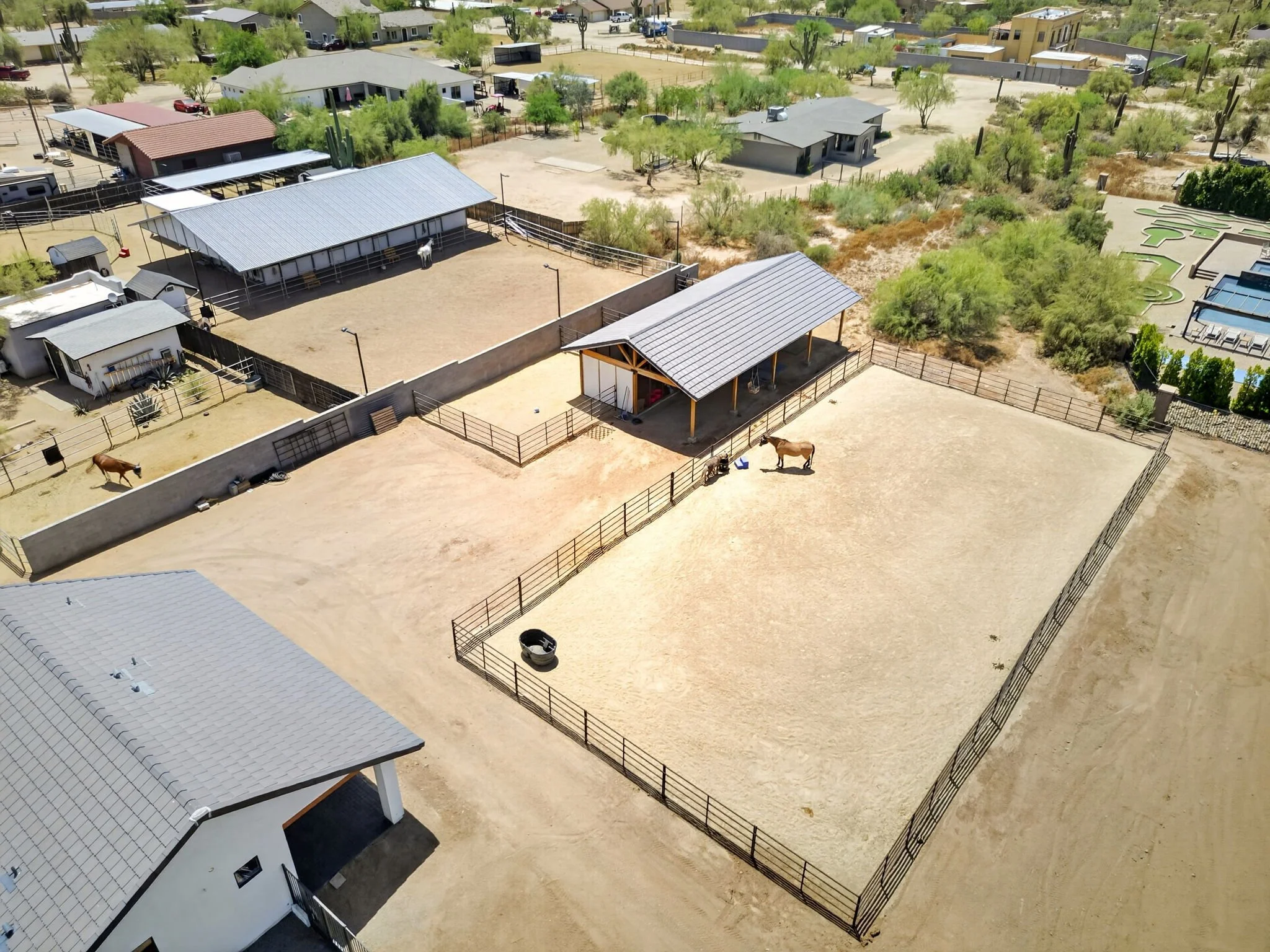 Aerial view of a ranch or farm with horses in fenced outdoor enclosures, green trees, and multiple small buildings with metal and shingle roofs, desert landscape in the background, and a swimming pool area.