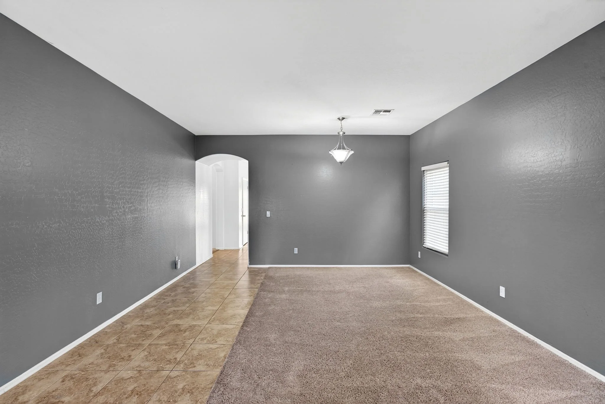 Empty living room with gray painted walls, tiled and carpeted floors, a chandelier, a window with blinds, and an archway leading to another room.