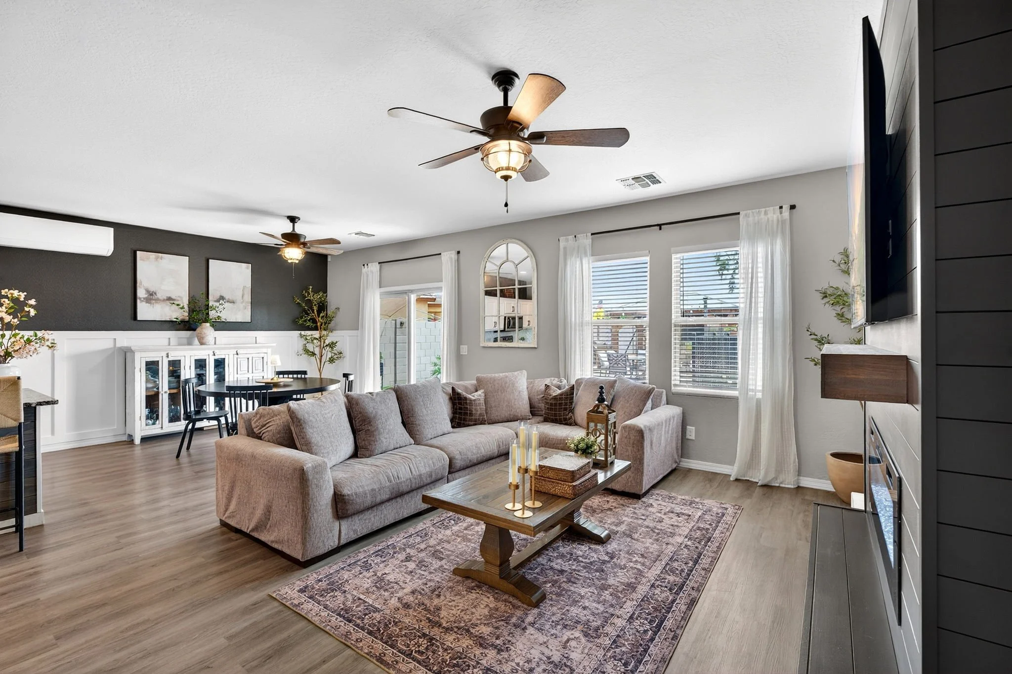 Living room with beige sectional sofa, wooden coffee table, large area rug, and multiple windows with white curtains. Ceiling fans and a dark accent wall with framed artwork are visible.