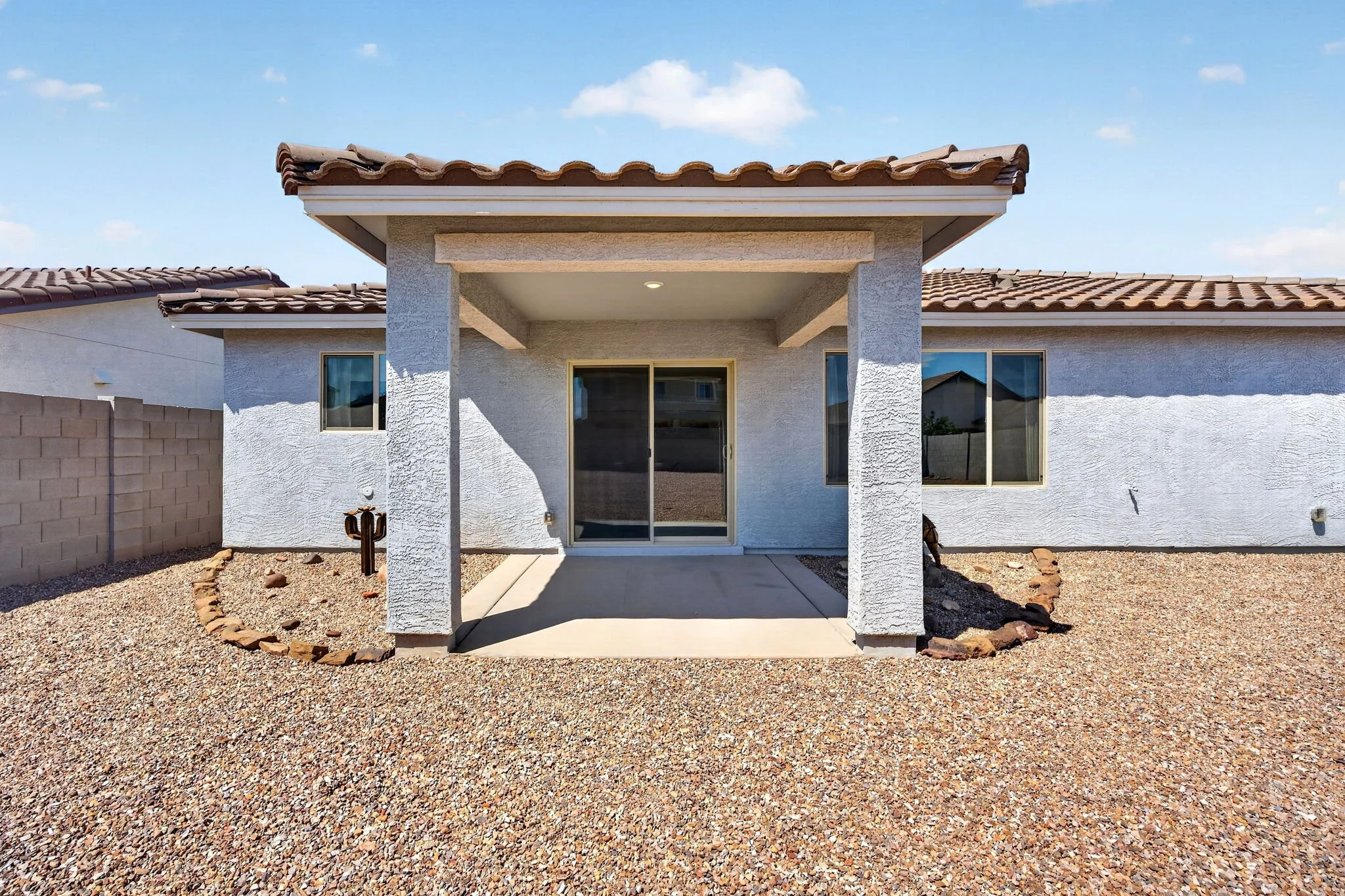 The back view of a white house with a covered patio, two windows, and a sliding glass door, surrounded by gravel yard with a circular rock border and a brick wall on the left side, under a blue sky with some clouds.