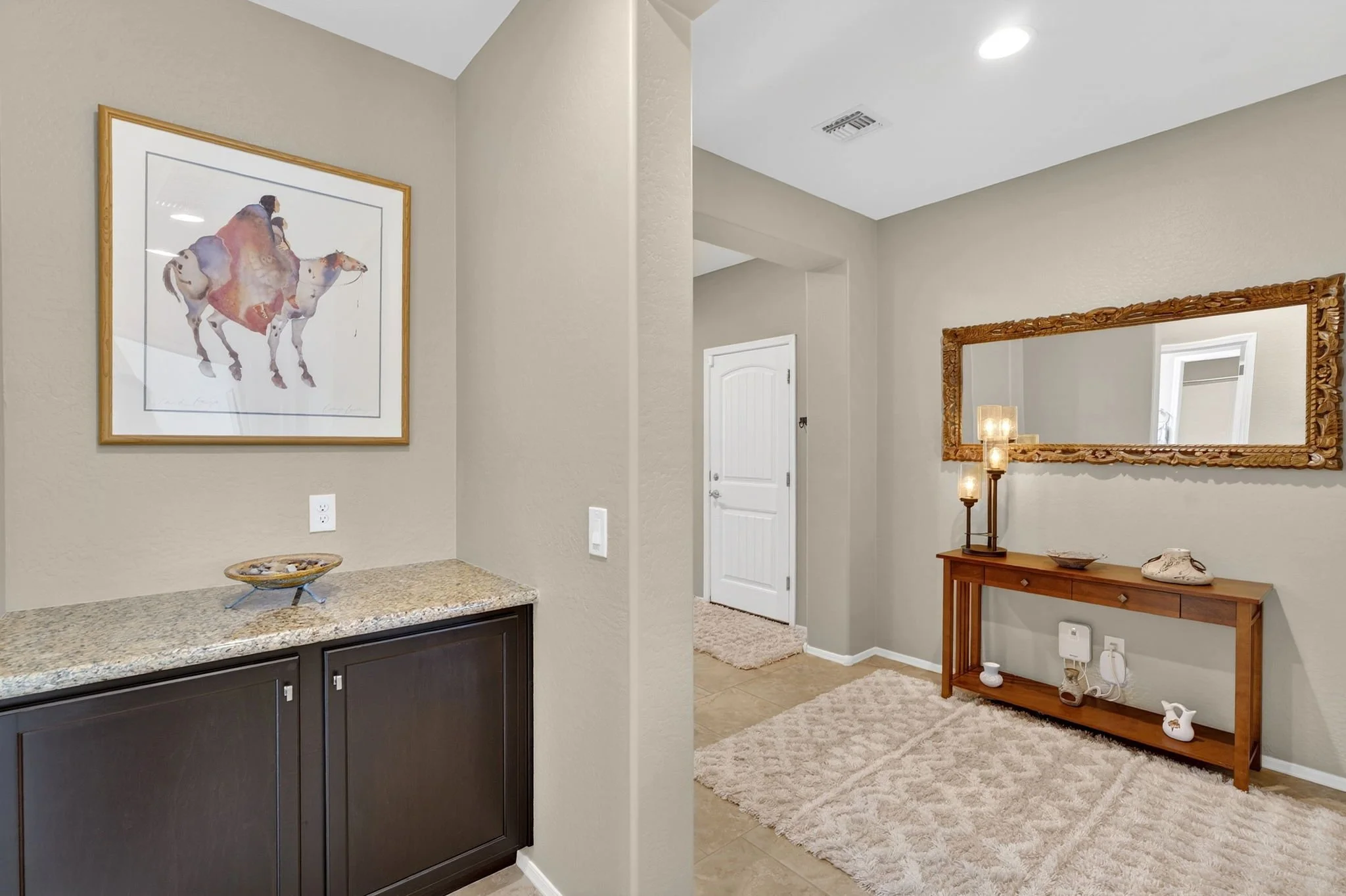 Interior view of a modern home entryway with beige walls, a large framed painting of a cowboy on a horse, a wooden console table with decorative items, a mirror with carved wooden frame, and a lamp with a beige shade.