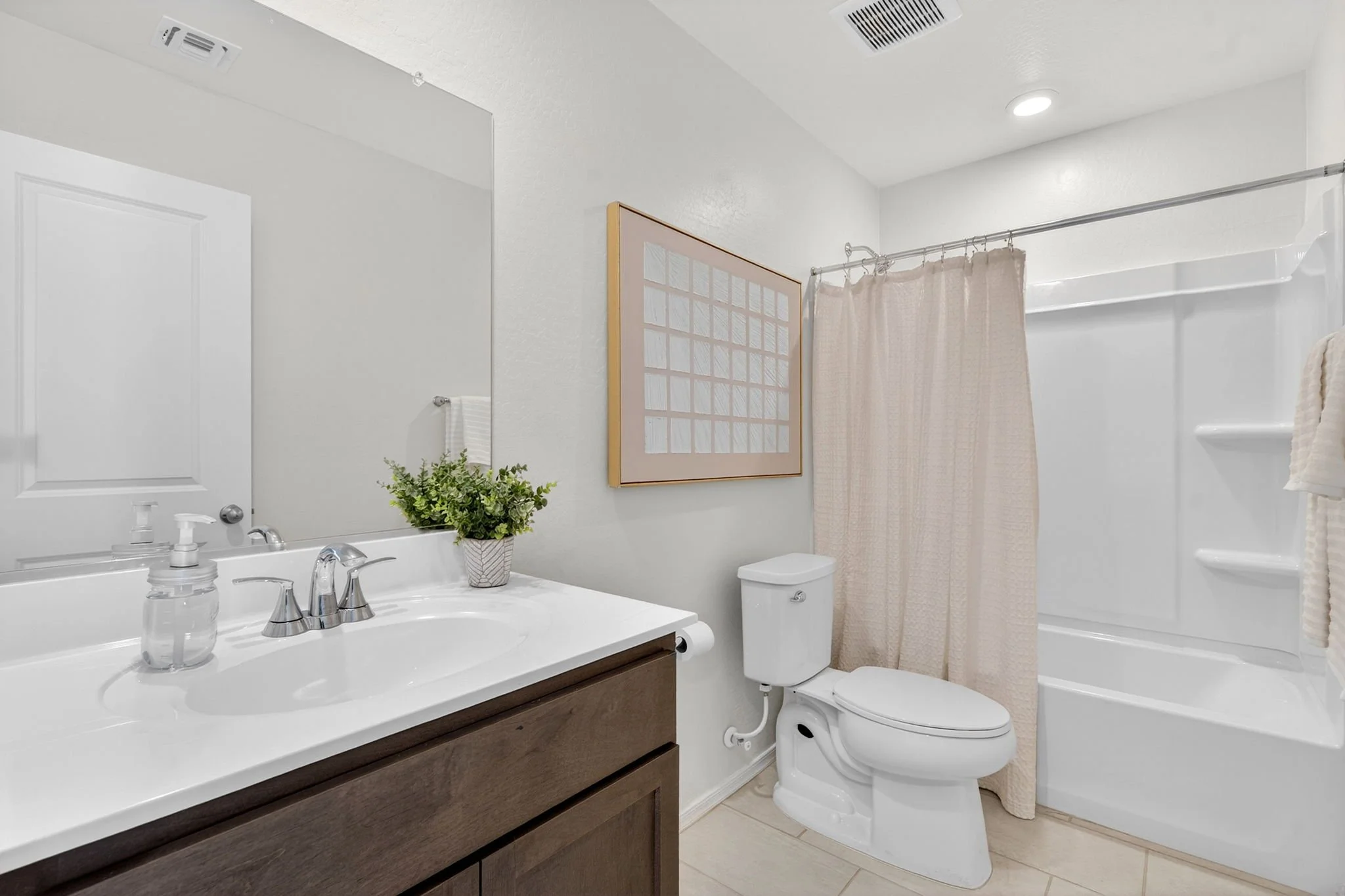 A clean bathroom with a white sink and a dark wooden vanity, a mirror above the sink, a white toilet, a floral plant in a white vase, a framed window, a shower with a beige shower curtain, and built-in white shelves in the shower area.