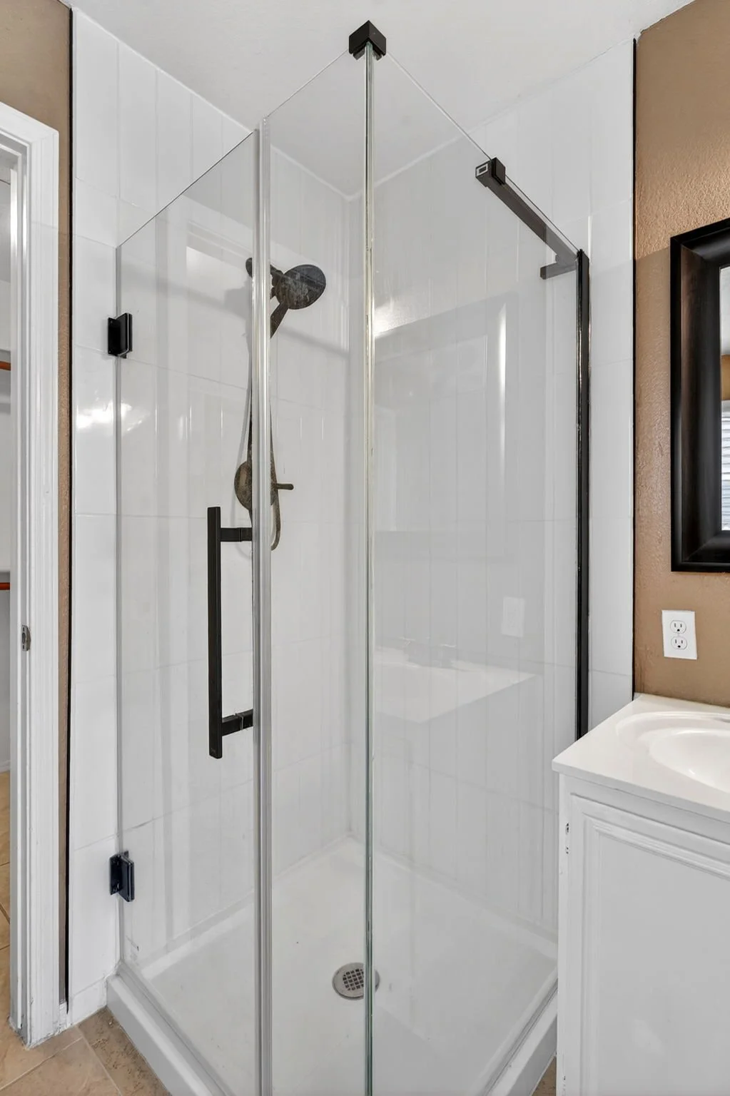 Glass shower enclosure with black metal hardware, white tiled walls, handheld shower head, and a white bathroom vanity with a sink next to it.