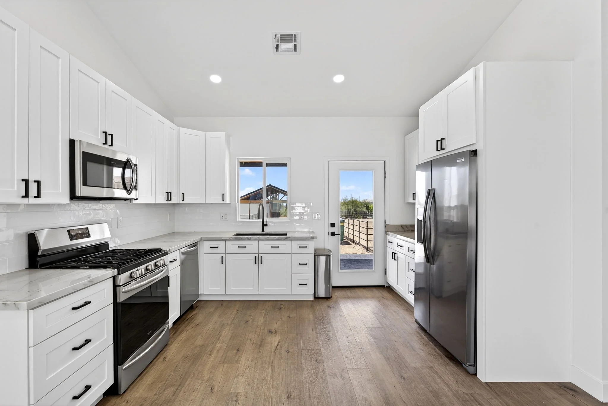 Modern white kitchen with stainless steel appliances, hardwood floors, and sunlight outside through windows and door.